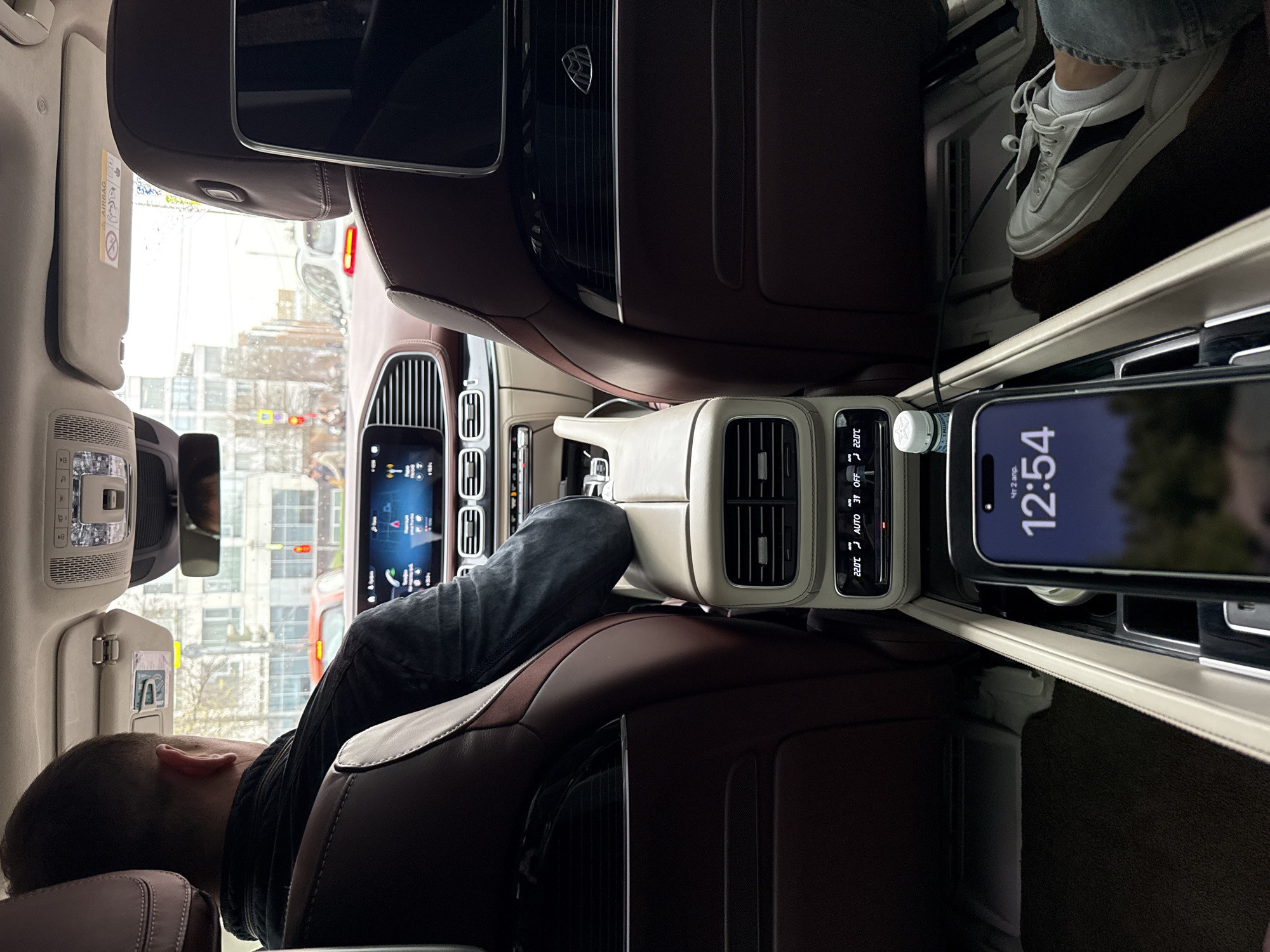 The image shows the interior of a luxury car, possibly a Maybach, from the rear passenger's perspective. The car is located in what appears to be a city, based on the view outside the window, which includes buildings and traffic signals. A person is reclining in one of the rear seats, with their head resting on the seat back. Another person's leg and foot, wearing a white sneaker, are visible in the other seat. A phone is placed in a charging slot, displaying a time of 12:54 and the date "Чт 2 апр". The climate control display shows a temperature setting of 22.0°C. The interior is a mix of dark brown and beige colors. Given the date format and the Cyrillic letters on the phone, it's likely the city is Moscow, Russia.