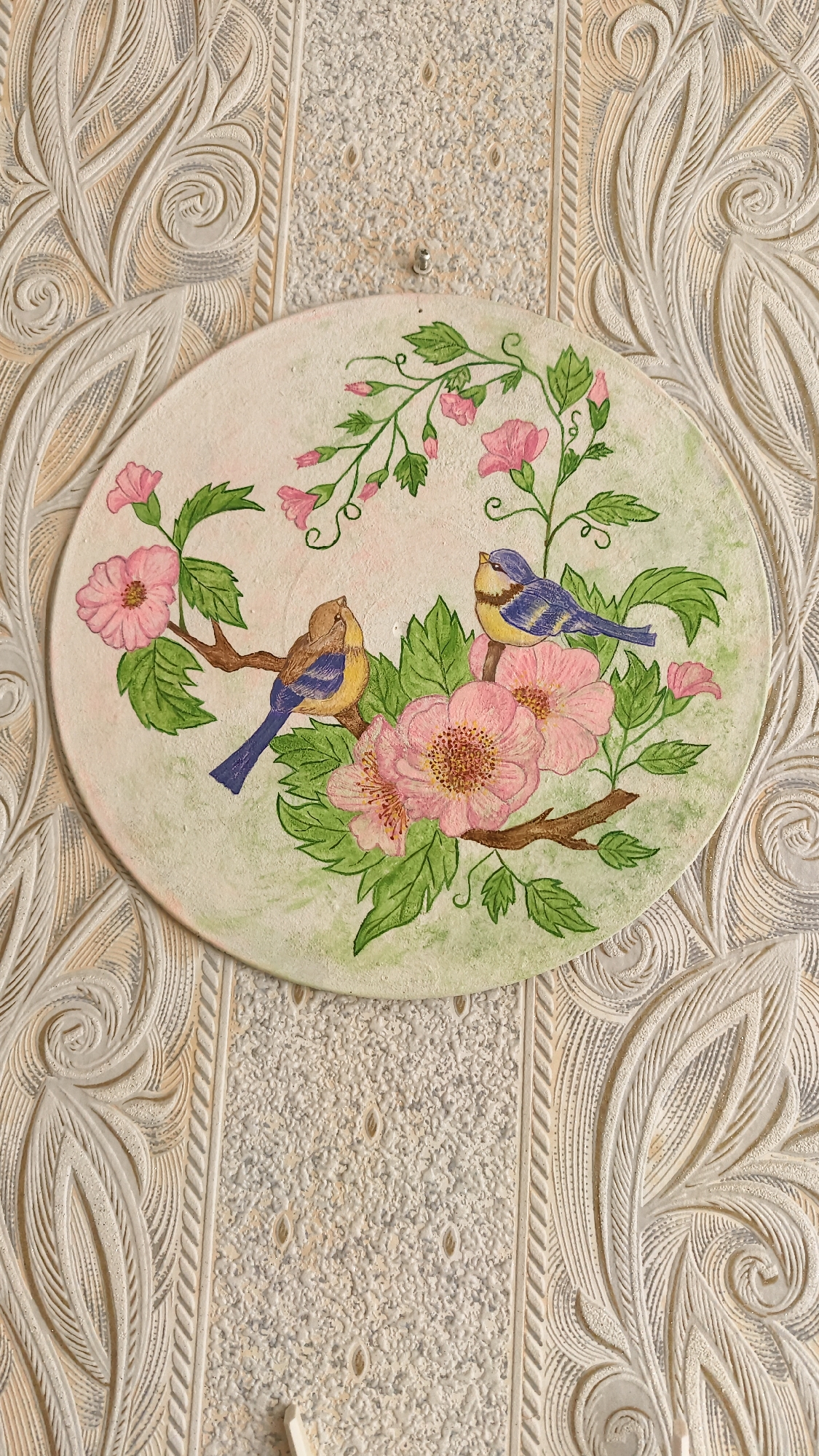 This media file features an oval-shaped decorative plaque depicting a nature scene, likely a piece of art or home decor. The plaque is painted with a light background, on which a branch of pink flowers and green leaves is depicted. Two birds are perched on the branch. One bird is primarily brown with blue and yellow accents, while the other is a vibrant blue and yellow. The artwork has a slightly textured, hand-painted feel.
The plaque is mounted on a wall with a distinctive textured wallpaper. The wallpaper has a repeating pattern of raised, decorative elements in shades of off-white and light beige, creating a visually rich backdrop. The textured wallpaper consists of two distinct styles: one with a finely granulated surface and small, raised oval shapes, and another with elaborate, raised scrollwork and leaf-like motifs. A small, silver hook is visible above the plaque, suggesting it is hanging on the wall.
The overall impression is one of a cozy, indoor setting, possibly a home in Horishni Plavni, Ukraine, given the location context. The lighting appears to be natural or soft artificial light, casting subtle shadows and highlighting the textures of both the artwork and the wallpaper. There is no visible text in the image. The scene is static, focusing on the decorative elements and their placement within an interior.