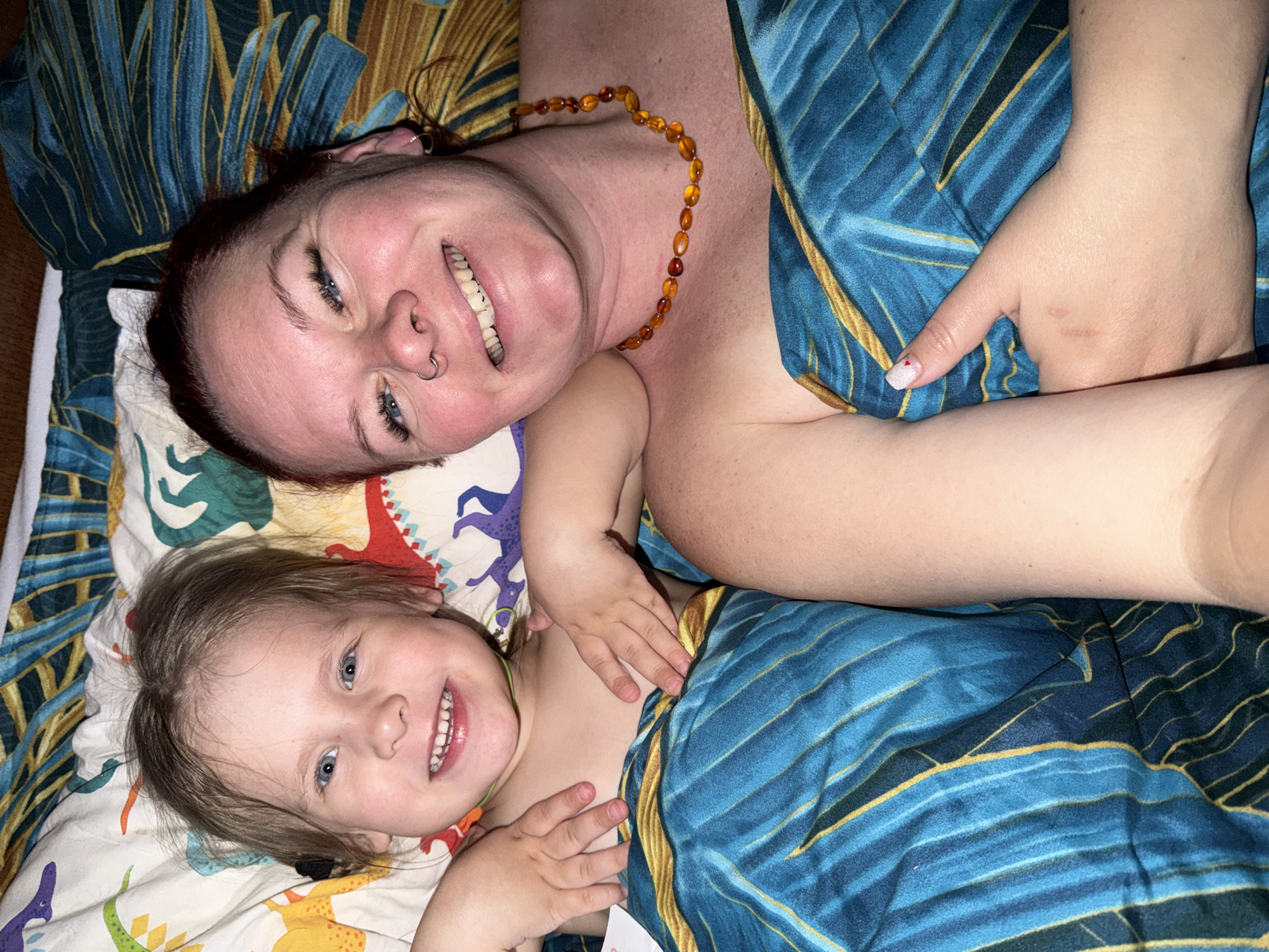 Two smiling people, likely a mother and child, are lying down next to each other. The child is on a dinosaur-printed fabric, and the adult woman has an amber necklace and lies on a patterned blue fabric. The location is not identifiable as Chaikovsky, Russia.