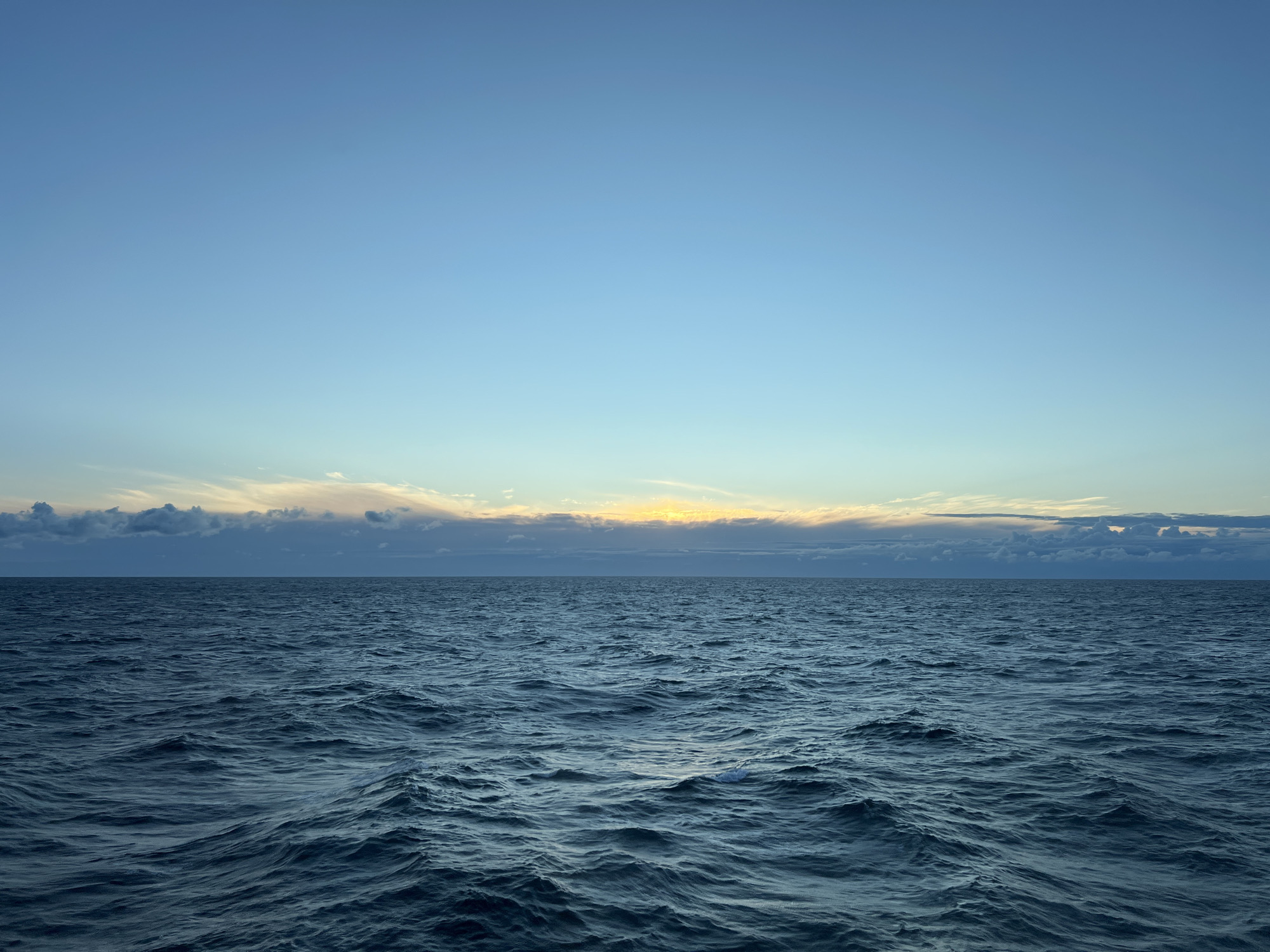The image depicts a vast expanse of dark blue ocean with moderate waves under a clear, pale blue sky. A band of clouds stretches across the horizon, tinged with yellow and orange, suggesting the presence of the sun just below or emerging from the horizon, possibly during sunrise or sunset. The scene is devoid of any human presence, structures, or landforms. The location is not identifiable as being within a city in Poland.