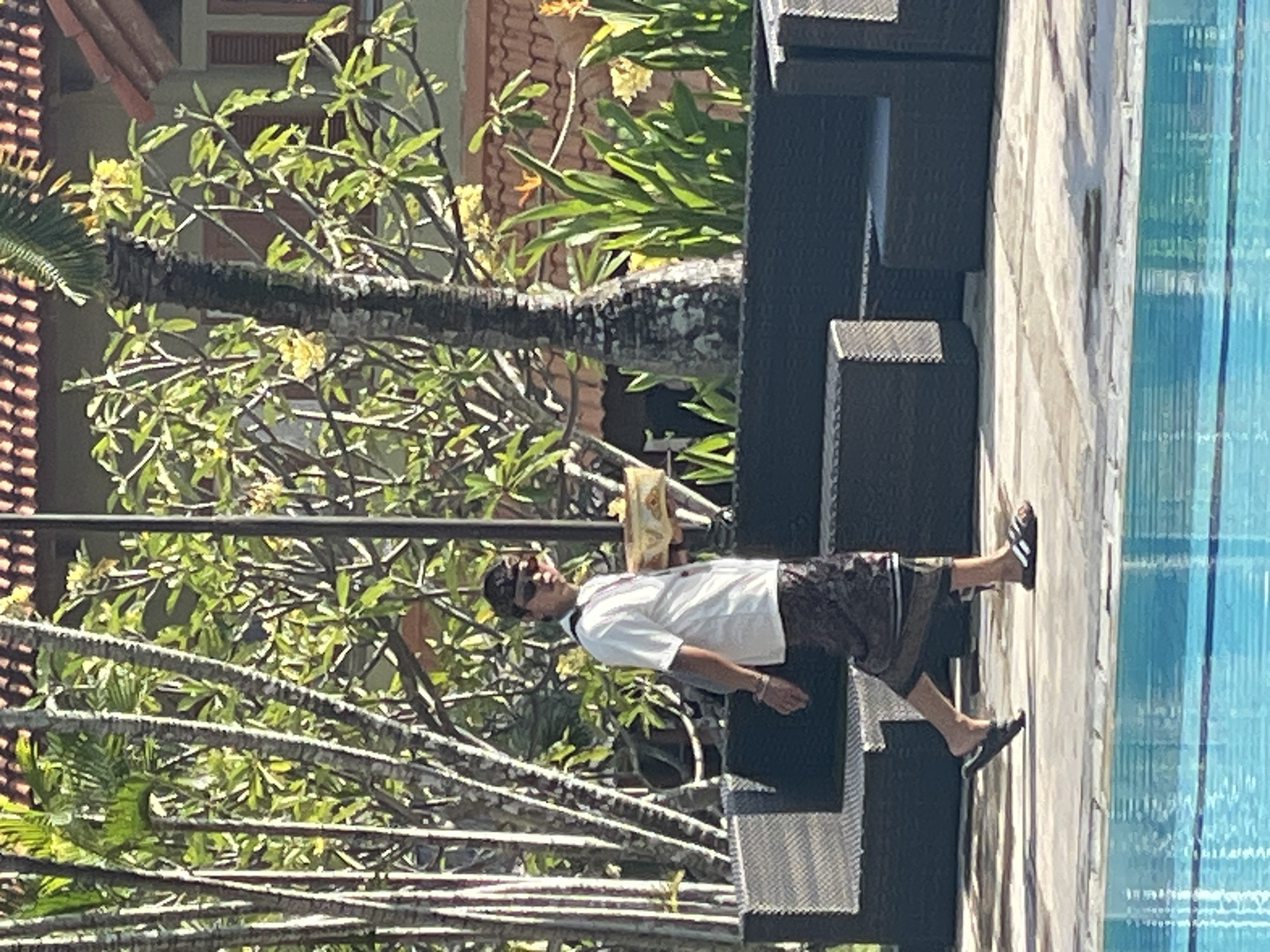 A person is walking on a tiled path next to a swimming pool in Kuta Selatan, Indonesia. The person is wearing a white t-shirt, dark patterned shorts, a dark head covering, and sunglasses. They are carrying a shallow, decorative bowl on their right hand. In the background, there are large, dark outdoor seating units and lush green foliage, including palm trees. The architecture in the background appears to be of a resort or hotel. The swimming pool in the foreground is filled with bright blue water.