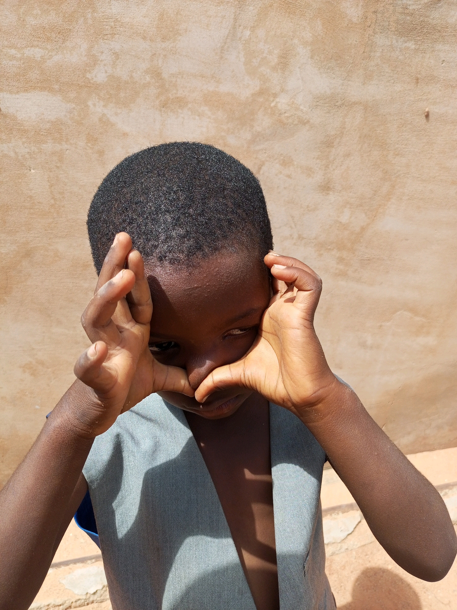 A young Black child, with dark curly hair and brown skin, is shown from the chest up, standing in front of a light brown, textured wall. The child's hands are raised to their face, with fingers framing their eyes in a gesture that could be interpreted as playful or secretive. The child is wearing a light grey vest over a blue garment. Sunlight casts a shadow on the child's chest and a portion of the wall. The ground is visible at the bottom of the frame, appearing to be a light-colored, rough surface. The image is an outdoor, daylight shot, likely taken in an urban environment given the wall and the clothing, suggesting it could be from Tempure, Nigeria.