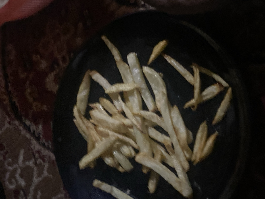 A black plate piled high with French fries dominates the scene. The fries are mostly pale yellow with hints of golden brown, appearing slightly greasy. The plate sits on a surface that is partially obscured, showing a textured, patterned material, possibly a rug or blanket, in dark browns and reds. The lighting is dim, casting shadows and creating a slightly muted tone.