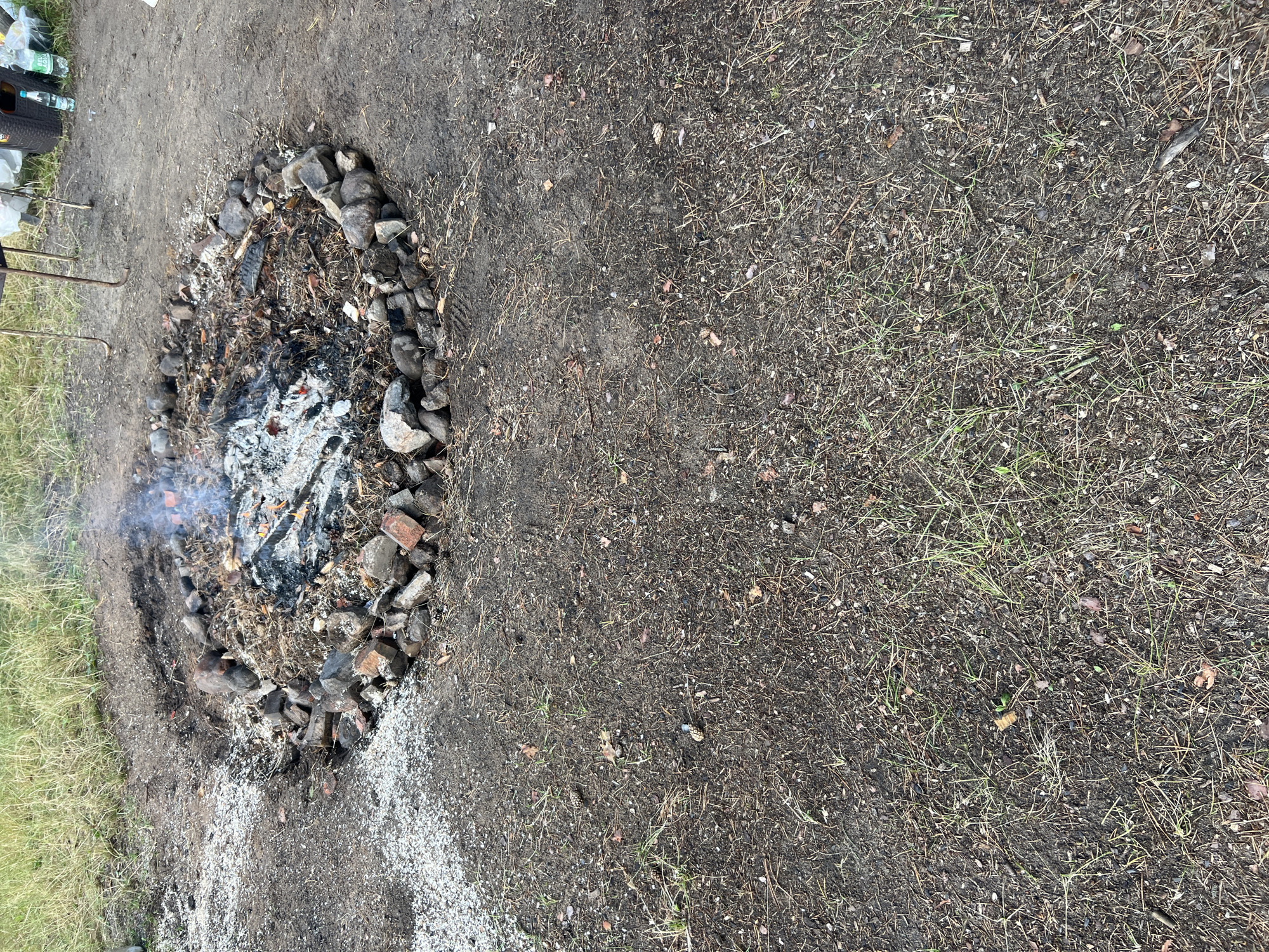 An outdoor, top-down view captures a fire pit constructed from stones and bricks in a campsite. The pit contains smoldering embers and ash, with wisps of smoke rising. Surrounding the fire pit is a mixture of dirt, sparse grass, pine needles, and small pebbles. In the upper right background, a metal grill, some plastic bags, and water bottles are visible. The ground is uneven and appears to be a natural, undeveloped area. The location is identified as Дражнаўскі сельскі Савет, Belarus.