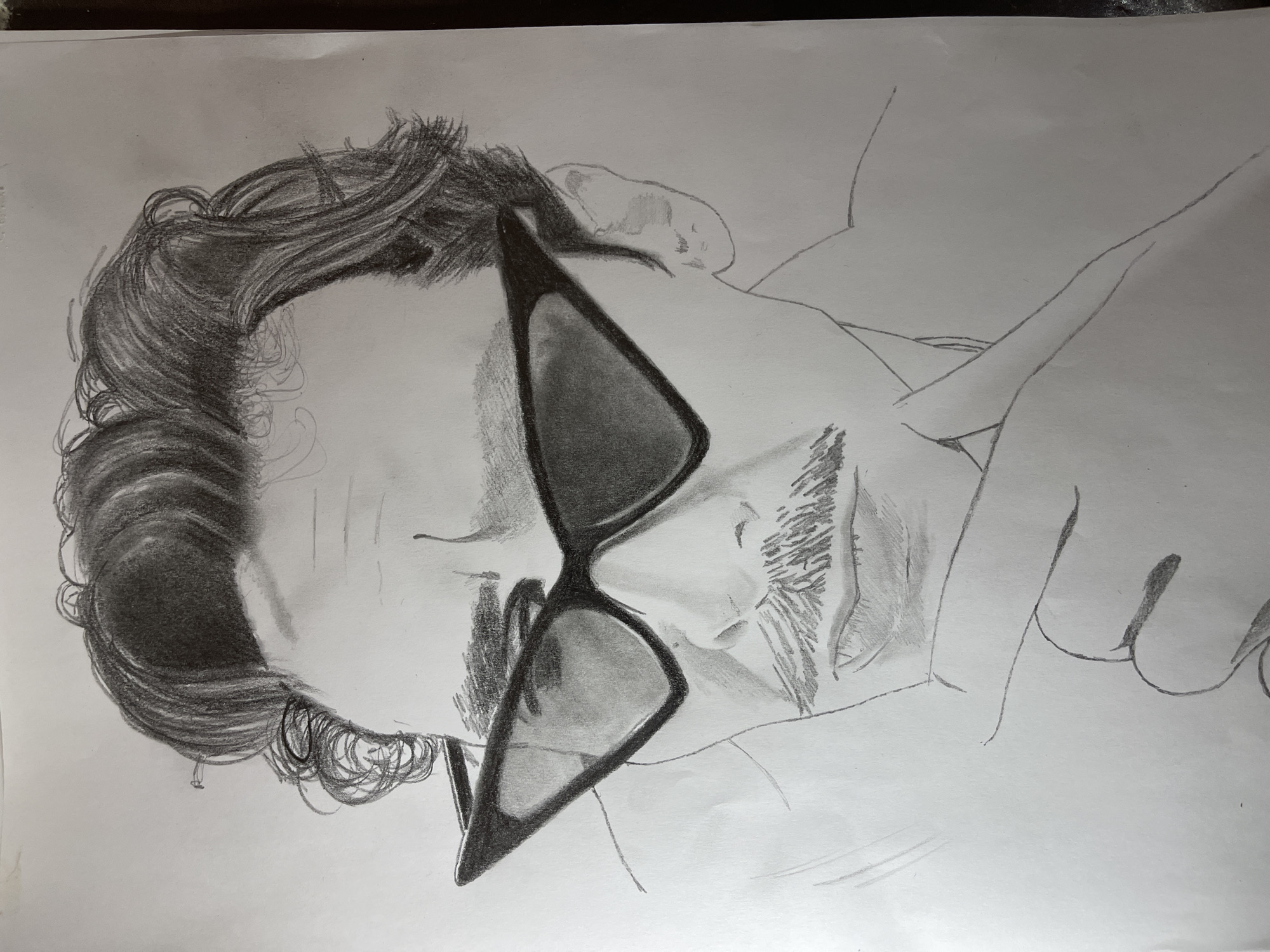 A pencil drawing shows a person's face wearing sunglasses and a partial view of the upper body. The person has dark, curly hair and a trimmed beard. The sunglasses have dark lenses. The drawing is on white paper. There is no discernible information to place the image in Jos, Nigeria.
