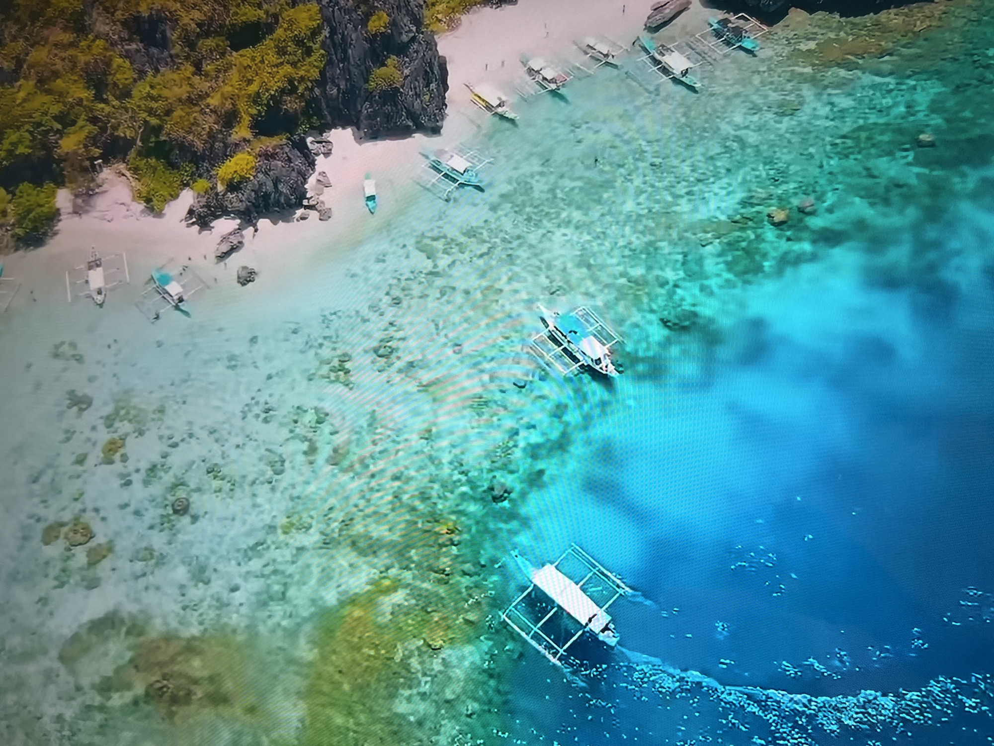 The image is an aerial shot of a tropical beach. A small island covered in vegetation is on the left side of the image. There is a sandy beach with several boats docked along the shoreline. The water transitions from shallow, clear turquoise near the beach to a deeper blue further out. Multiple boats with outriggers are visible in the water. The seabed is visible in the shallow areas, showing light-colored sand and rocks. The location is not in Basingstoke and Deane, United Kingdom; it is a tropical coastal environment.