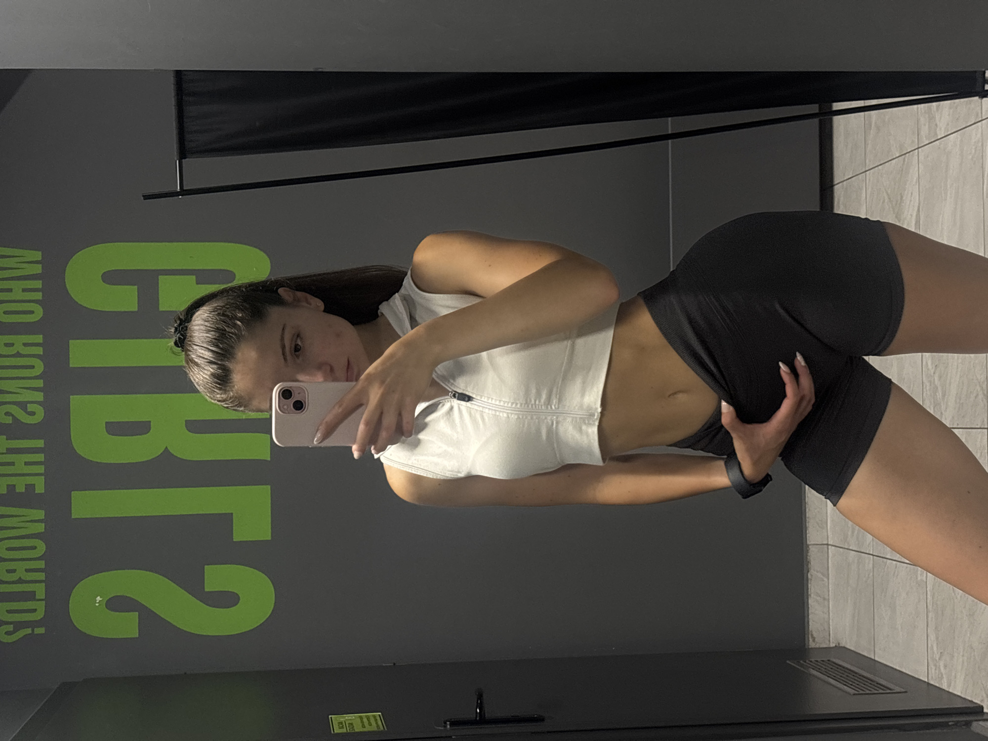 In a gym locker room in Varna, Bulgaria, a young woman with dark hair pulled into a ponytail stands in front of a mirror, taking a selfie. She is wearing a white zip-up crop top and black athletic shorts. Her left hand is placed on her hip, just above her shorts. Behind her, a large green graphic on the wall reads "GIRLS WHO RUN THE WORLD?". The floor is tiled, and a black curtain hangs on the right side of the frame. A vent is visible on the gray wall to the left.
