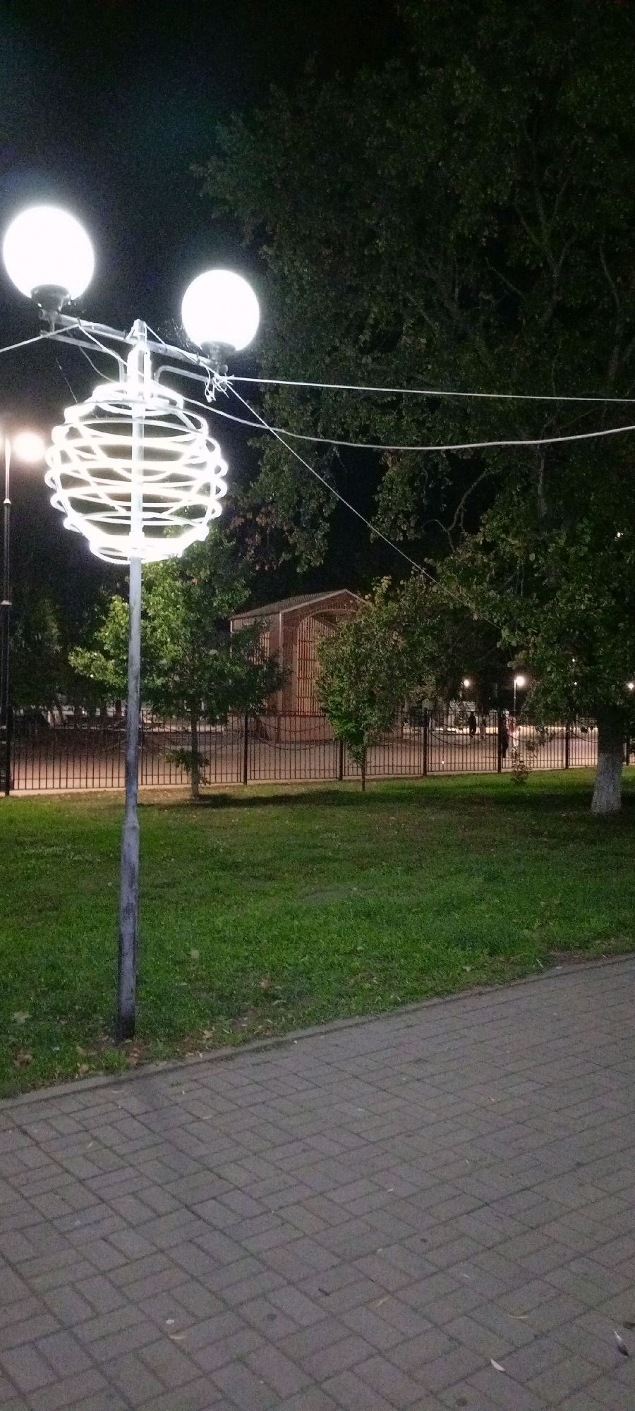 An exterior, eye-level, nighttime shot features a decorative lamp post in the foreground, on the left side of the image, illuminated with a bright, white glow. The lamp post itself is silver-colored, and extends upwards with a complex, circular, white light fixture resembling a spherical nest of wires. Above the circular fixture are two spherical lamps, also glowing white. The lamp post stands on a paved walkway composed of grey, rectangular bricks laid in a basket-weave pattern.
To the right of the walkway is a patch of green grass, appearing well-maintained. Beyond the grass, a black metal fence runs horizontally across the frame, separating the park area from what appears to be a building in the background. This building has a wooden facade and an arched entrance, suggesting it might be a pavilion or a small structure within a park.
Behind the fence, several trees with dark green foliage are visible, with their branches and leaves creating shadows against the night sky. In the distance, through the fence and trees, faint lights can be seen, and silhouettes of a few individuals suggest a public park or urban space is being utilized at night. The overall scene conveys a sense of a public park at dusk or night, possibly in the city of Kirsanov, Russia, judging by the context provided. The lighting creates a somewhat dramatic and atmospheric ambiance.