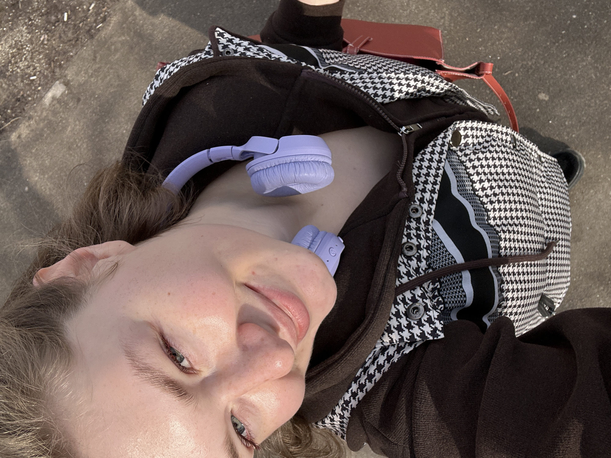 A close-up, upward-facing selfie shows a young woman with fair skin and light brown hair. She is smiling slightly, with a hint of lip gloss on her lips. She is wearing a dark brown hooded jacket, partially zipped open, revealing a houndstooth patterned garment underneath. A pair of light purple headphones rests around her neck. A glimpse of a red leather bag strap is visible in the upper portion of the frame. The background appears to be a concrete surface, possibly a sidewalk. There are no other visible details to indicate the location within Kazan, Russia.