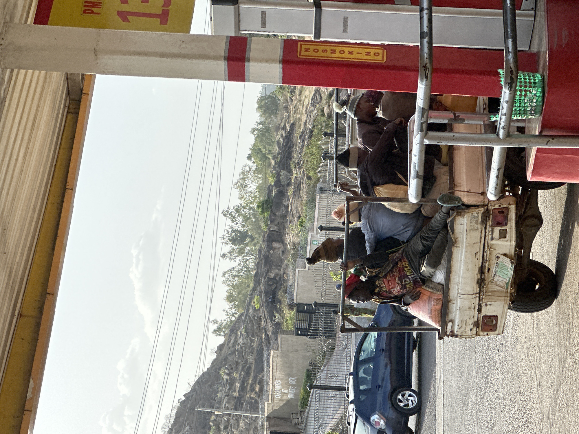 Captured outdoors in the city of Jos, Nigeria, the image shows a passenger-laden vehicle pulled over to the side of the road near what seems to be a gas station. The passengers are dark skinned. On the left, a blue car is parked, and in the background, there’s a mountain or hillside. A “NO SMOKING” sign is visible on the vehicle. The image appears to have been rotated counter clockwise.