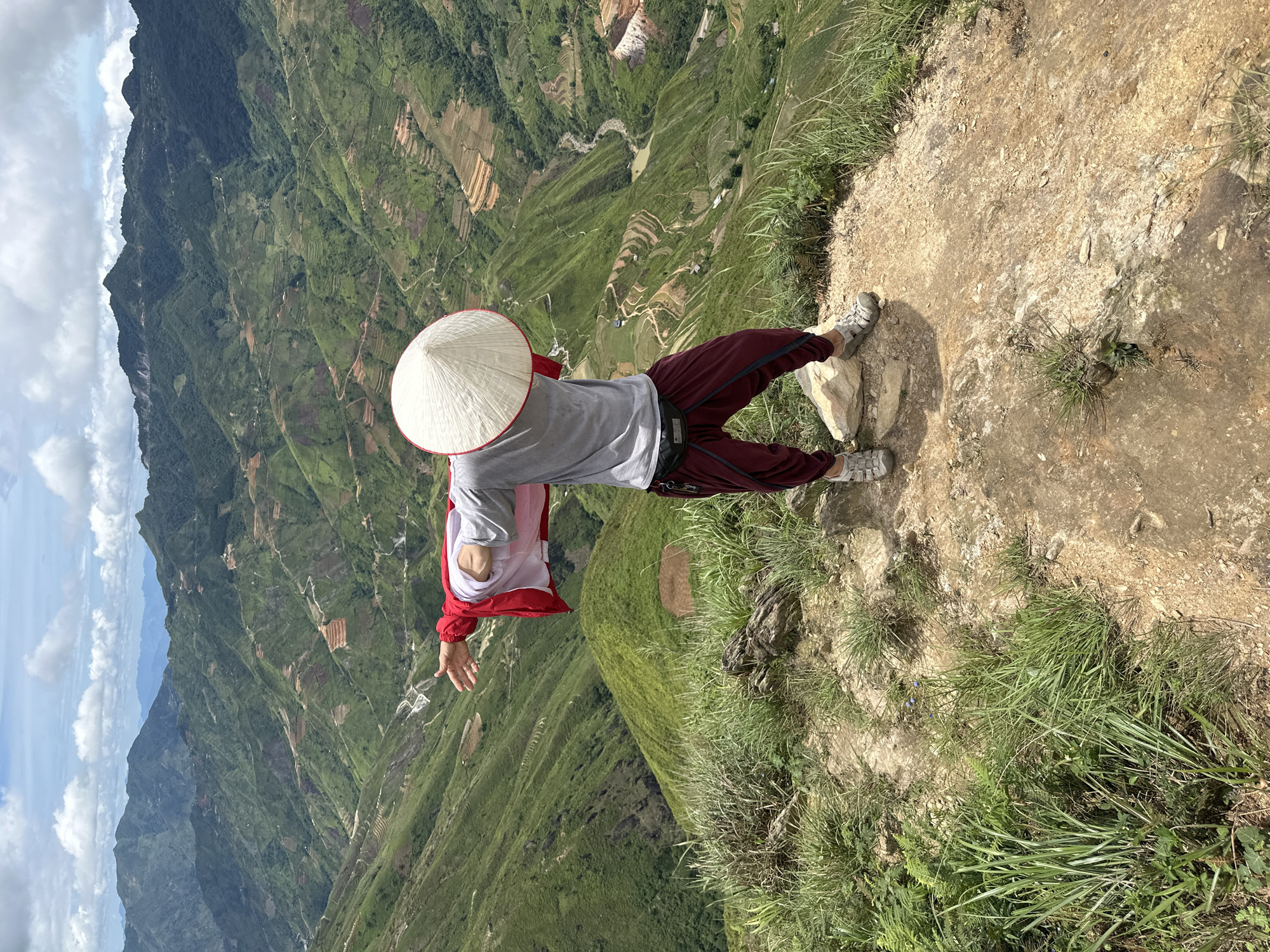 In the image, a person stands on a rocky, grassy precipice overlooking a vast, verdant valley in Ta Xua, Vietnam. The person is viewed from behind, wearing a traditional conical hat, a grey t-shirt, a red jacket draped over their shoulders, and maroon pants. Their arms are outstretched, as if embracing the expansive landscape. The valley below is characterized by rolling, green hills dotted with agricultural terraces, winding roads, and a meandering river. The sky above is a mix of blue with scattered white clouds. The scene evokes a sense of awe and connection with nature.