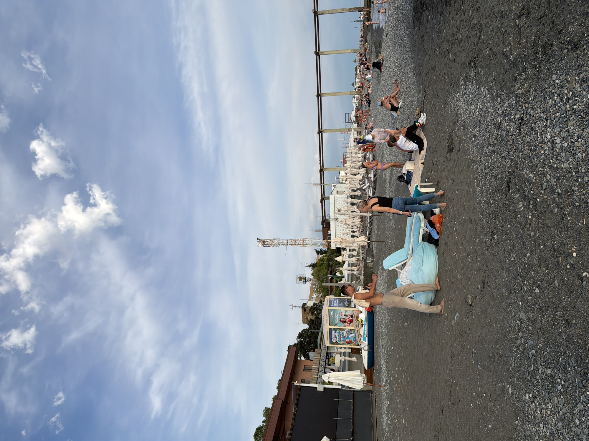 The image depicts a pebbly beach in Sochi, Russia, under a partly cloudy sky. In the foreground, a man wearing tan pants and no shirt stands on the beach, his towel draped over his shoulder. He is near a large, light blue beanbag chair. Further back, a woman in a black tank top and blue jeans stands next to the beanbag. Other people are scattered across the beach, some sitting, some standing, and some walking.
The background reveals a row of beach umbrellas and lounge chairs, suggesting a developed recreational area. Buildings with reddish-brown roofs are visible behind the umbrellas, and a tall communication tower stands further inland. A blue and white watercraft is positioned on the beach near the buildings. The overall scene is one of a typical summer day at the beach.