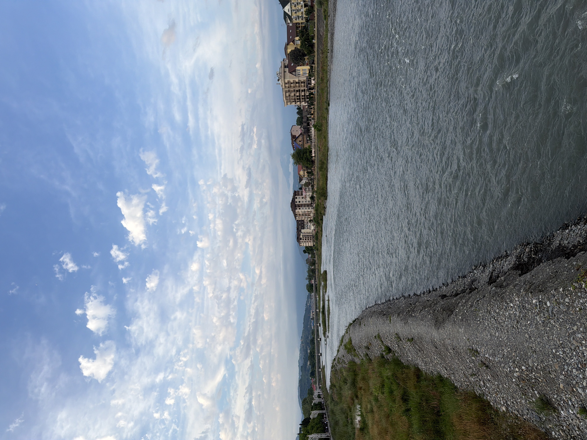 This is an eye-level, landscape, full shot of a wide, grey, slow-moving river in Sochi, Russia, likely in the early evening. On the left bank of the river, there is a raised embankment covered in grey pebbles and dirt, with dry grass and some green vegetation growing on it. Beyond the embankment, there are some buildings, a bridge, and trees. On the opposite bank, a line of multi-story residential buildings, some with balconies and a few with tiled roofs, stretches along the river. The sky above is a pale blue with scattered white, wispy clouds. The overall impression is a serene, urban riverside scene.