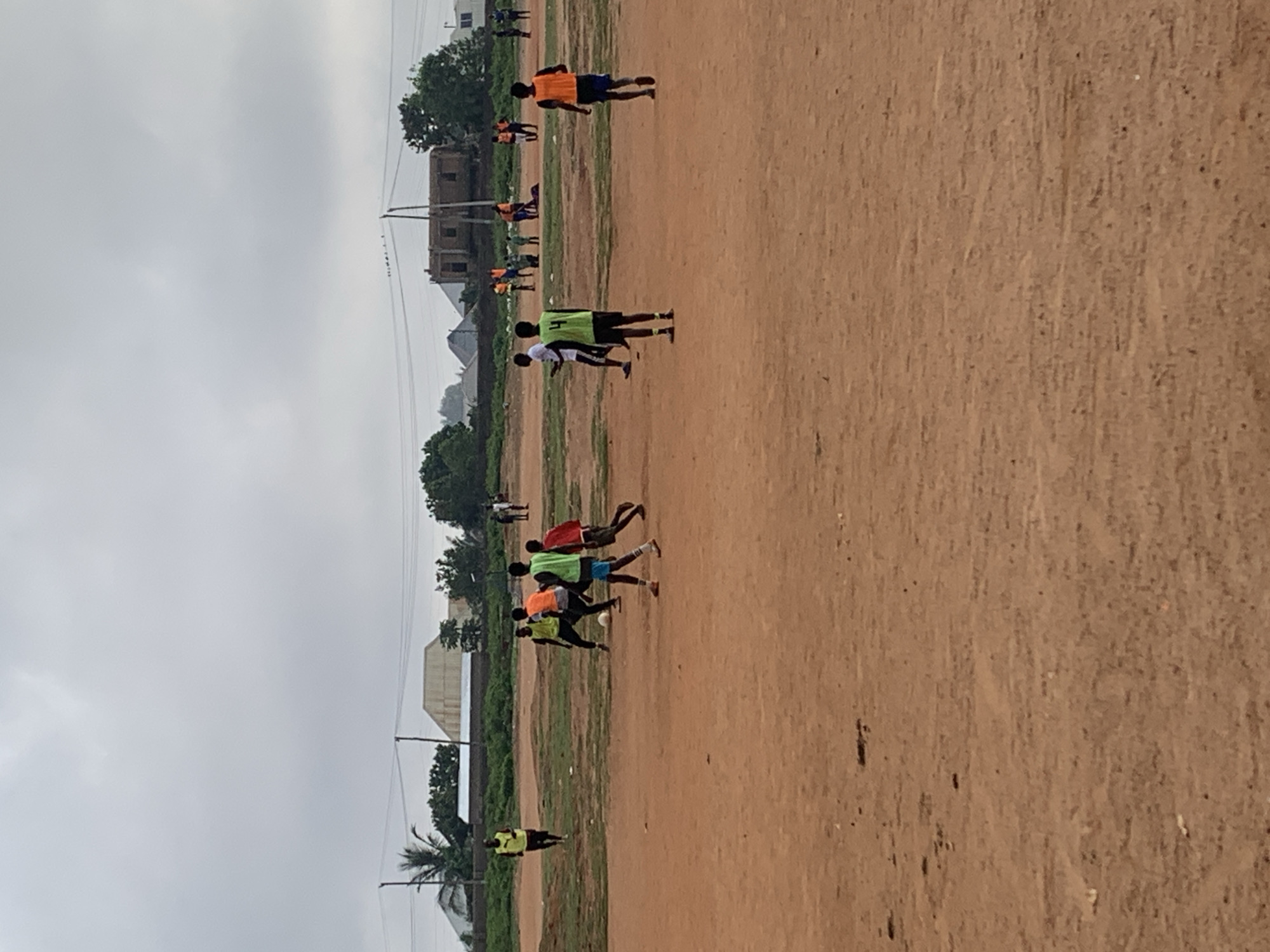 In Bukuru, Nigeria, on a dusty, reddish-brown soccer field, a group of young men in brightly colored vests are engaged in a soccer game. The sky is overcast with heavy gray clouds. In the background, a few buildings with corrugated metal roofs and some trees are visible. The overall scene depicts an informal soccer match taking place on a rural or unpaved field under a cloudy sky.
