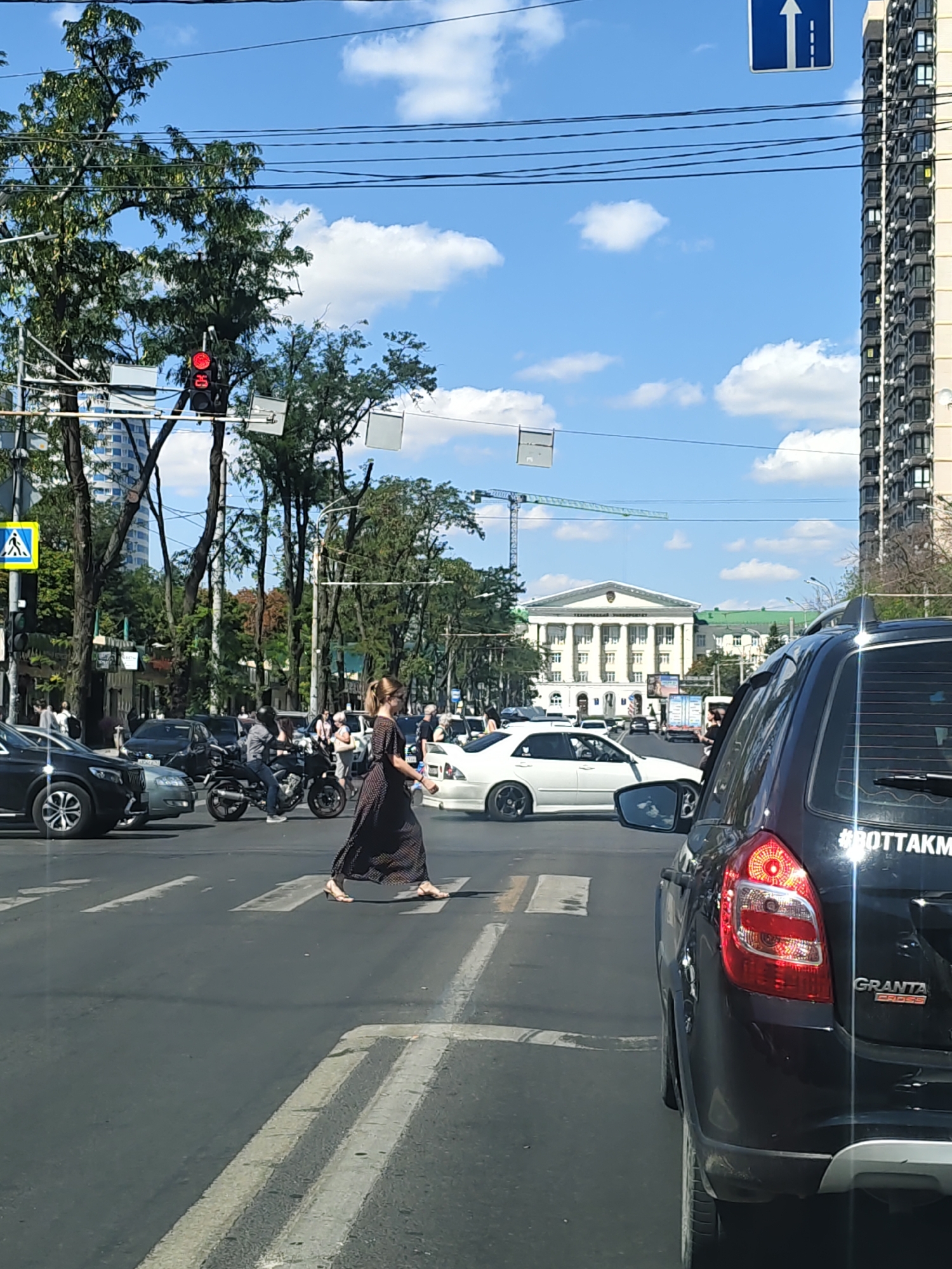 On a sunny day in Rostov-on-Don, Russia, a street scene unfolds at an intersection. The camera is positioned inside a black LADA Granta Cross, looking forward. In the foreground, the rear of the LADA is visible. Across the intersection, a woman in a long, dark patterned dress is walking across a pedestrian crossing. She is mid-stride, with her right leg forward and her left leg trailing. To her left, a white sedan is stopped at the intersection. Behind the woman, a motorcycle with two riders is also present.
The intersection itself is marked with white pedestrian crossing lines. Traffic lights are visible; the one closest to the viewer appears to be red. A pedestrian crossing sign with a yellow background and a white symbol of a person walking is also visible on the left side of the intersection.
In the background, several trees line the street. Further back, a large, white classical-style building with columns and a grand facade dominates the skyline. A construction crane is also visible in the distance, suggesting ongoing development. On the far right, a tall, modern apartment building with many windows can be seen. The sky is blue with scattered white clouds. Overhead, power lines are strung across the scene.
