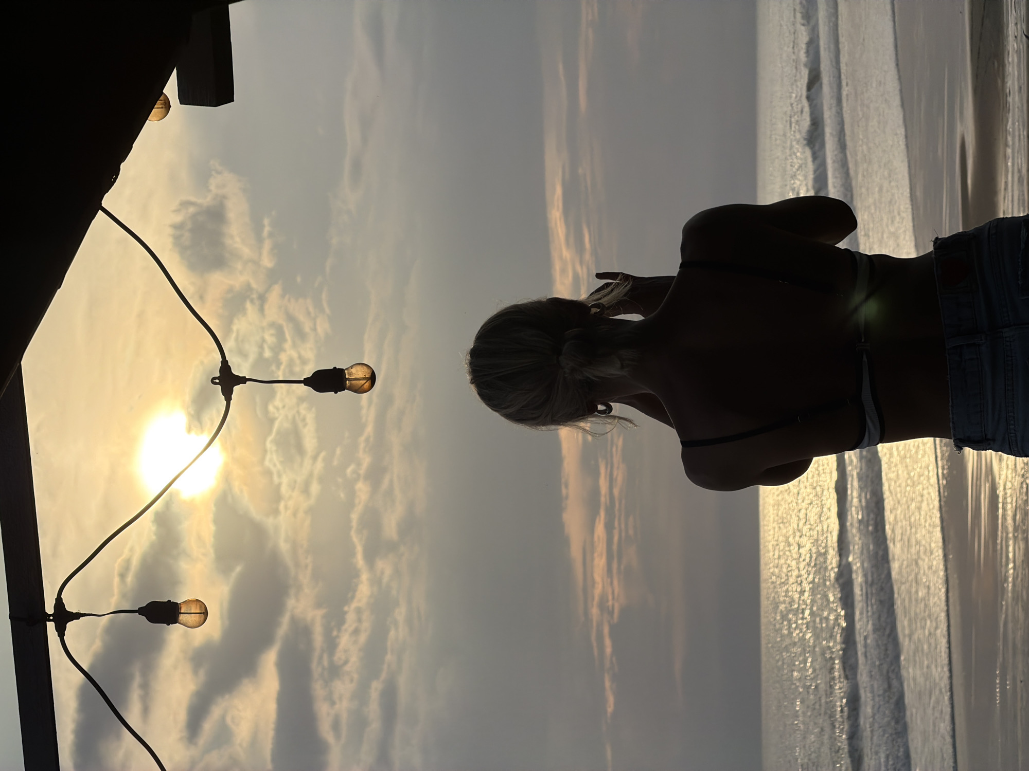 The image captures a beach scene at sunset, with a silhouette of a woman in the foreground, facing the ocean. She appears to be wearing a bikini top and shorts. Above her and to the left, string lights are suspended from a dark wooden structure, with the setting sun visible between them. The ocean and beach stretch out into the distance, reflecting the light of the sunset. It is not possible to ascertain where, exactly, this photo was taken in Kediri, Indonesia from just the image.