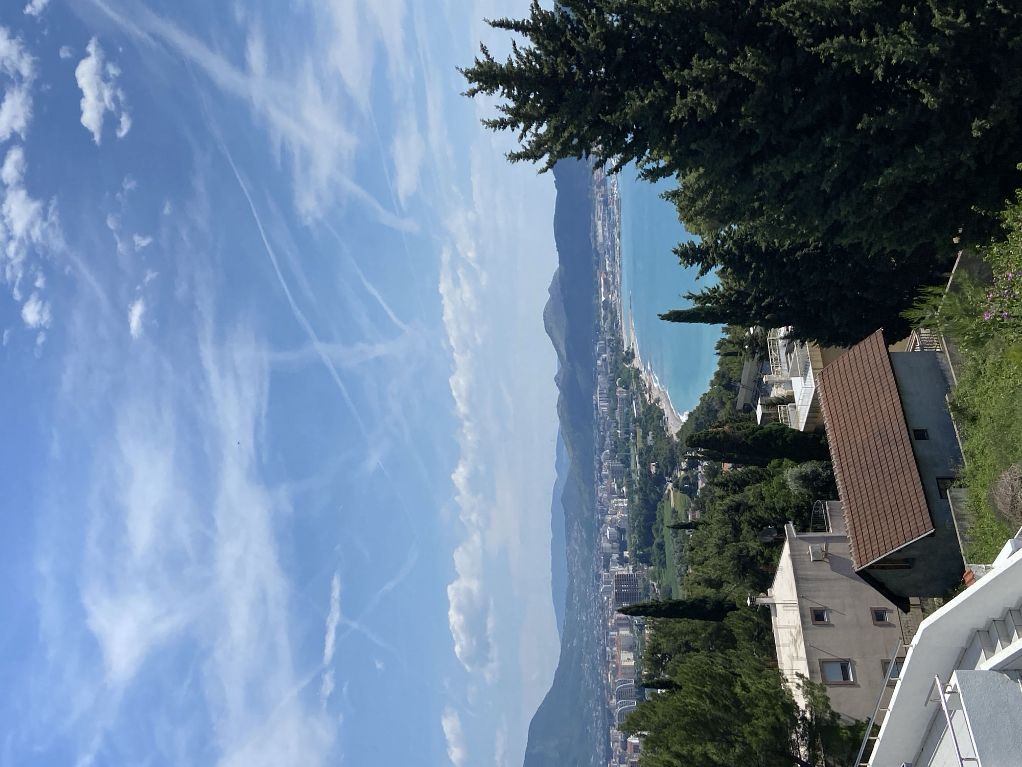The image presents a scenic view of Šušanj, Montenegro, from an elevated position. The foreground includes rooftops of buildings with brown and white exteriors and a staircase leading up to the elevated viewpoint. The middle ground features a panoramic view of the coastline, including the turquoise waters of the Adriatic Sea, sandy beaches, and the urban landscape of Šušanj. Mountains rise in the background, partially obscured by clouds and haze. The sky is blue with scattered white clouds and contrails. Evergreen trees are prominently featured in the right side of the frame, adding a natural element to the urban and coastal scenery.