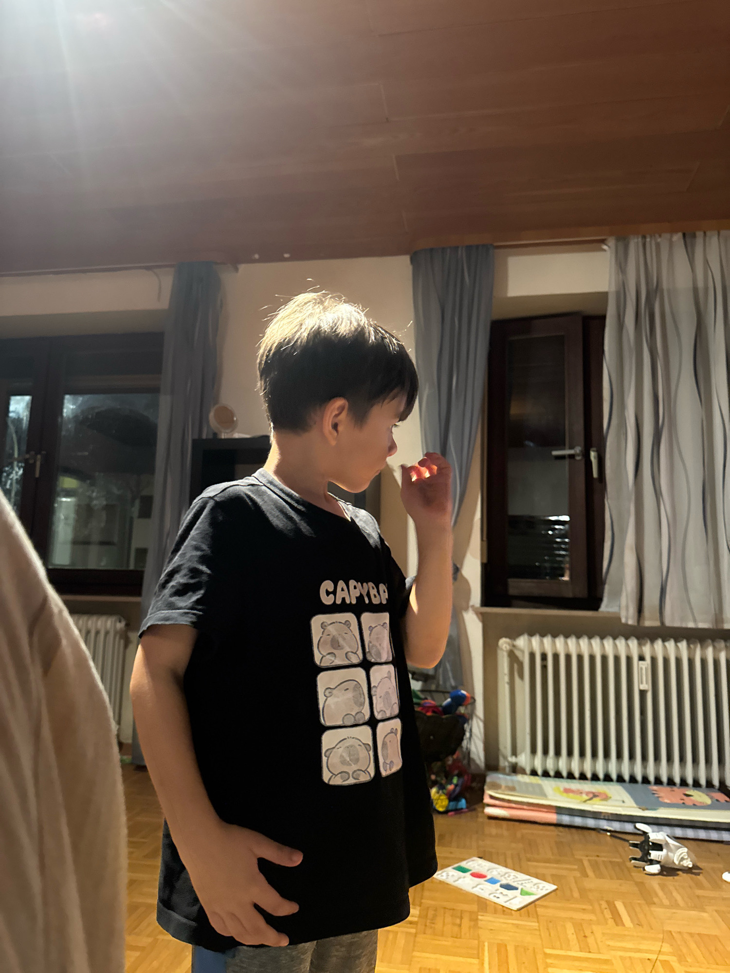 The image depicts a young boy standing indoors, likely in a residential setting. He is wearing a black t-shirt with a graphic print of several cartoonish bears in boxes and the text "CAPYBAR". His right hand is raised to his face, near his mouth. The background shows a wooden ceiling, curtains covering windows, a radiator, and toys scattered on a wooden floor. The lighting suggests it is either evening or a dimly lit room. There is no explicit indication that this location is in Überherrn, Germany, nor any contextual cues within the image to support such a claim.
