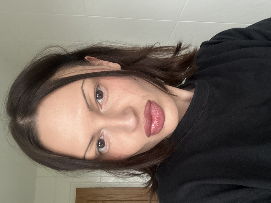A female individual with dark brown, medium-length hair styled with a center part is depicted in a close-up, slightly high-angle selfie. She has dark brown eyes, thin, drawn eyebrows, and prominent cheekbones with blush applied. Her full lips are glossy and pursed. She wears a plain black crew-neck sweatshirt, and a small stud earring is visible in the upper cartilage of her right ear. The background indicates an indoor setting, featuring a light brown wooden door on the left and white ceramic tiles with visible grout lines on the right. The scene is located in the city of Tunbridge Wells, United Kingdom.