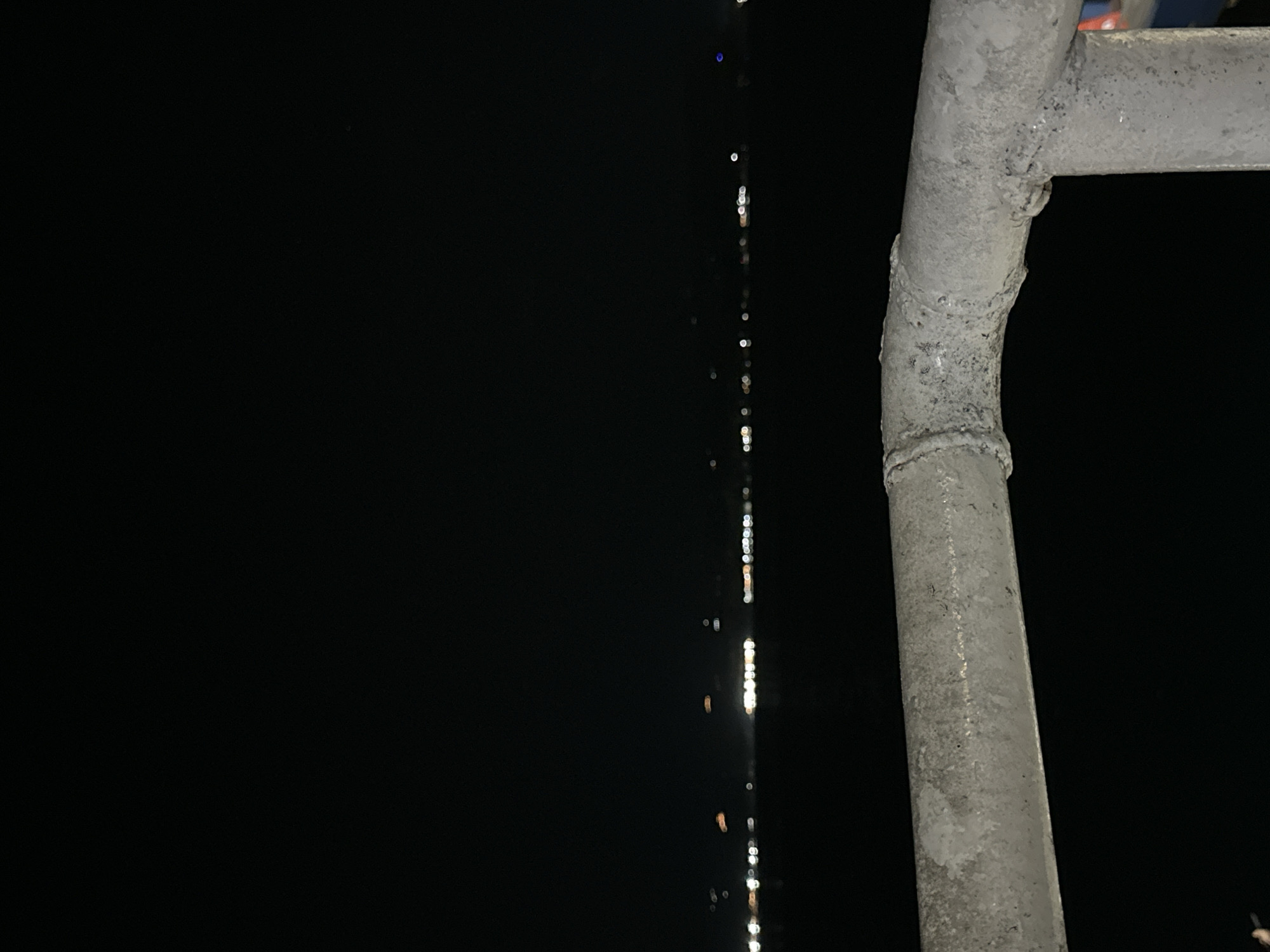 The image presents a nighttime scene with a prominent, weathered metal structure on the right side, likely part of a railing or scaffold. This structure is composed of dull gray, possibly galvanized or painted pipes, showing visible welding seams and some surface imperfections like corrosion or dirt. The background is overwhelmingly dark, a deep black, indicating late evening or night.
Running vertically through the central left portion of the black background is a line of small, blurry, and bright light sources. These lights vary in intensity and color, appearing as bokeh, suggesting they are distant or out of focus. They could be reflections on water, streetlights, or lights from buildings in the distance.
There are no people or discernible activities taking place. The composition focuses on the contrast between the textured metallic foreground and the dark, subtly lit background. No text is visible in the image. The overall impression is one of stillness and urban night, with artificial lights piercing the darkness. While the image is set in Davao City, Philippines, there are no specific visual cues within the photograph itself to identify this location.