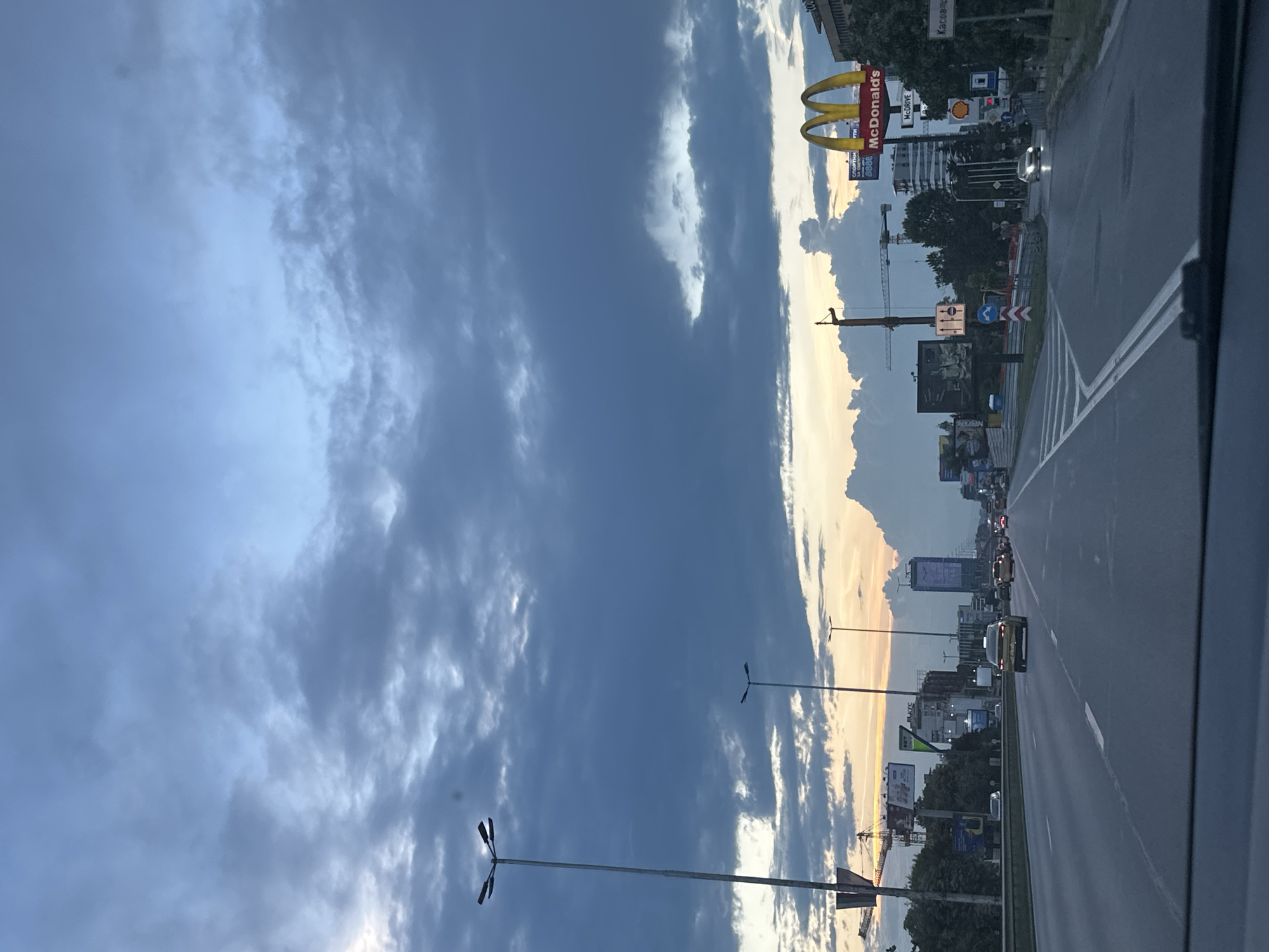 The image depicts a wide, multi-lane highway with traffic in Sofia, Bulgaria, under a dramatic, cloudy sky. The perspective is from inside a vehicle, suggesting a passenger or driver's view. The sky is a dominant feature, filled with dark, brooding clouds that are beginning to break apart, allowing some soft, golden light from the setting sun to filter through, creating a contrast between the dark and light elements.
Along the right side of the highway, there are several commercial signs and structures. Most prominently, a large McDonald's sign with its iconic golden arches is visible. Below it, there is a sign for "McDrive." Further down the road, a Shell gas station sign can be seen. Other billboards and advertisements are scattered along the roadside, some featuring images and text that are partially obscured by distance and the angle of the shot.
On the left side of the highway, a tall street lamp with multiple lights stands out. In the distance, several buildings and construction cranes indicate urban development and city infrastructure. A tall, modern building with a digital display or clock face is visible further down the highway, contributing to the urban skyline.
The highway itself has clear lane markings and appears to be a major thoroughfare. Several vehicles are on the road, including cars of various colors and models, moving in the same direction as the viewer. The presence of traffic lights and road signs suggests a regulated traffic environment. The overall scene captures a moment on a busy highway as evening approaches, characterized by the interplay of urban elements, vehicular movement, and a dynamic sky.