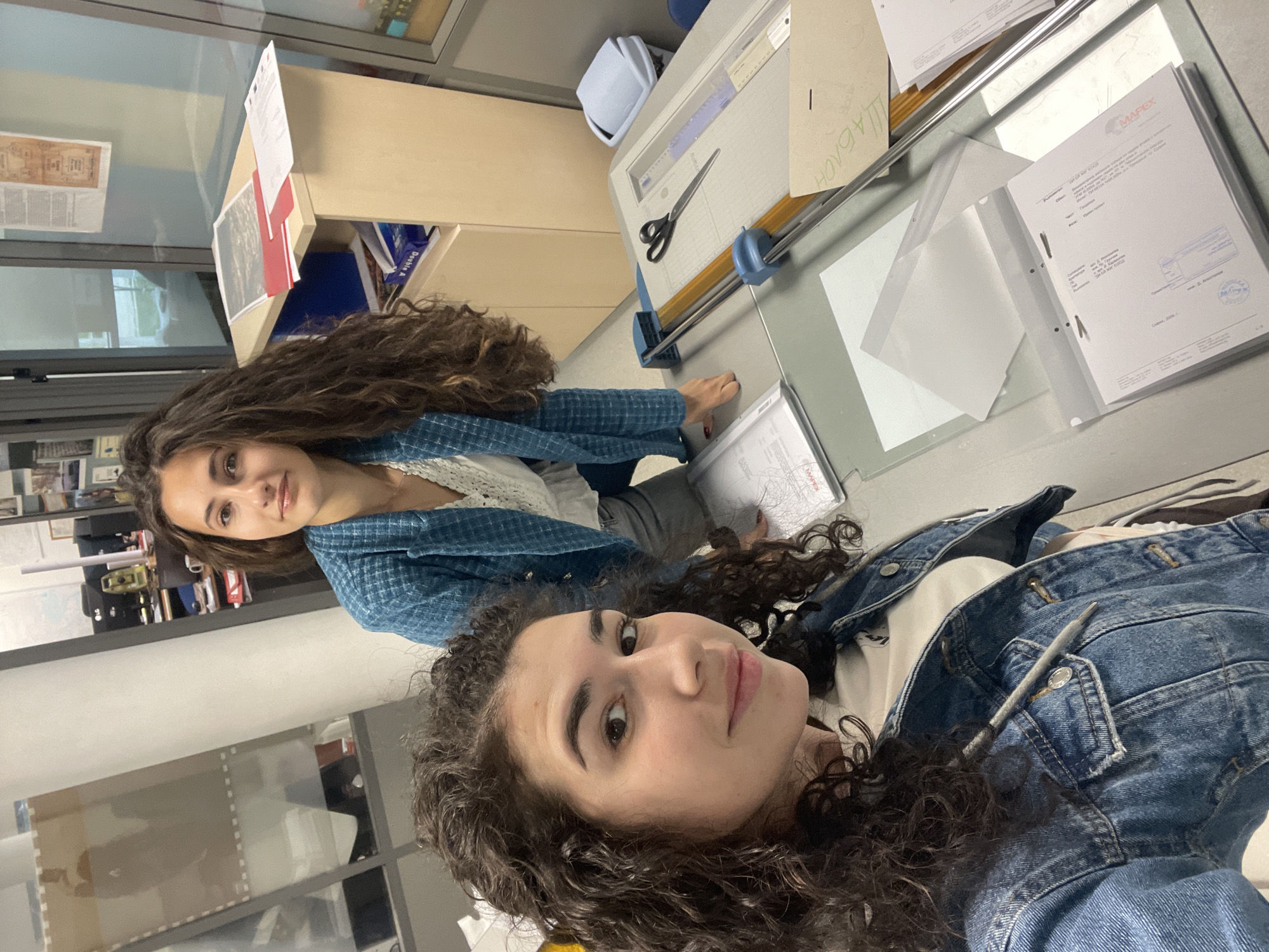 Two young women with curly brown hair pose in what appears to be an office or studio space. One woman wears a denim jacket and has a pen tucked into its pocket. The other woman wears a blue and white checkered top. They are positioned around a large table with various documents and office supplies, including a paper cutter, scissors, and ring binder containing paperwork. The environment suggests an academic or professional setting, potentially related to design or architecture. I cannot determine the location of Sofia, Bulgaria, from the image.