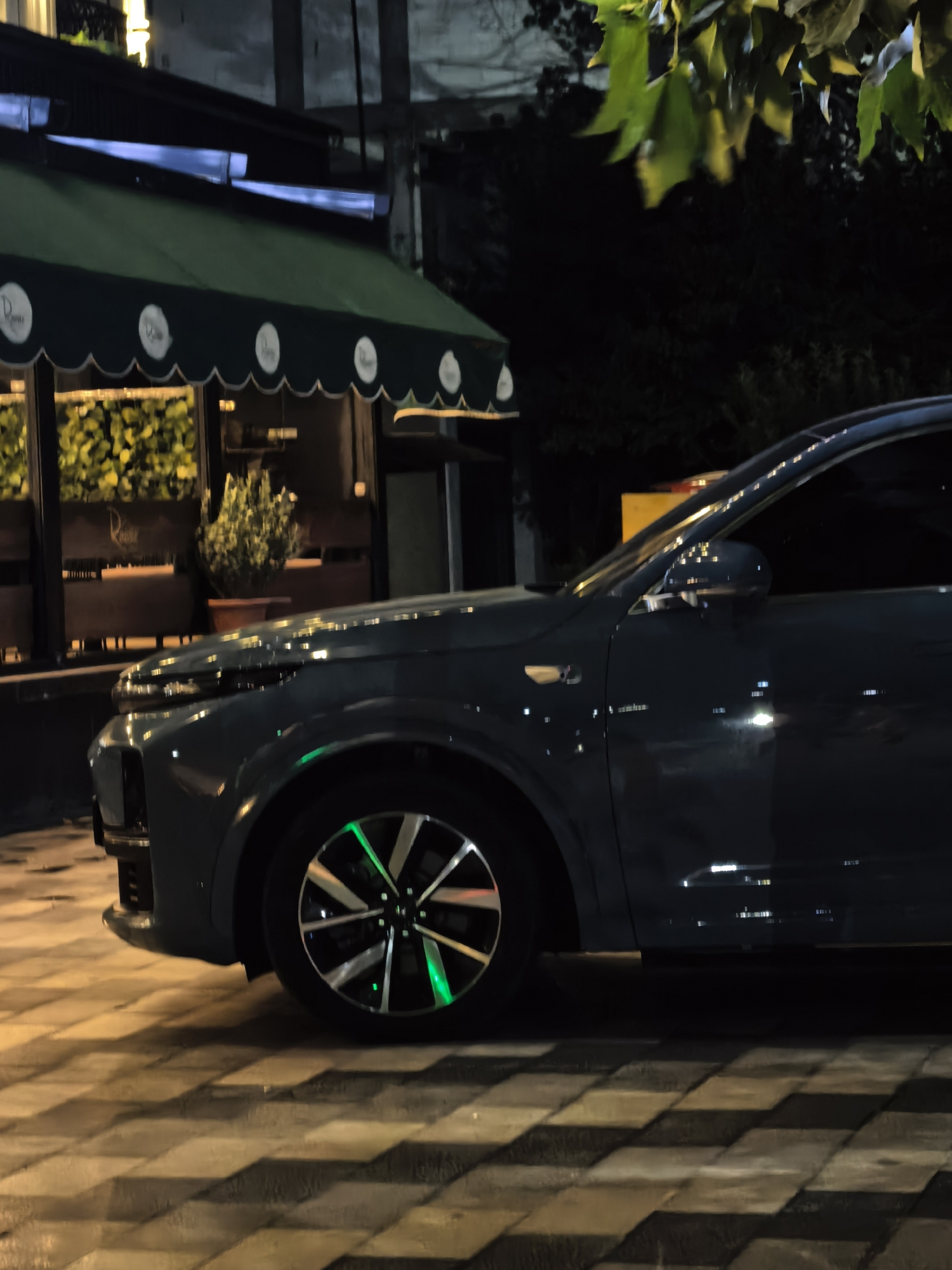 A dark-colored SUV is parked on a patterned pavement street at night. The car has a distinctive wheel design with green accents visible. In the background, there is a building with a green awning, likely a cafe or restaurant, with interior lighting illuminating potted plants and signage. Trees with green leaves are visible to the right, partially obscuring the sky. The overall scene suggests an urban setting at night. This location is identified as Fergana City, Uzbekistan.