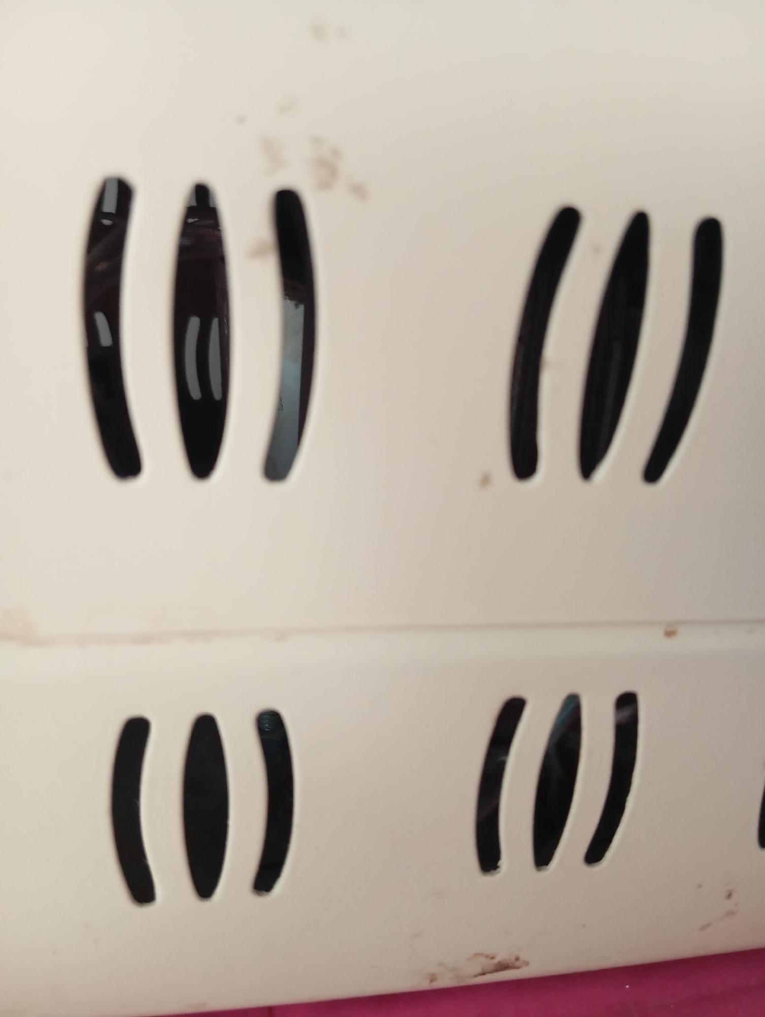 The image shows a close-up of a white surface with multiple parallel slots. The slots are oval-shaped and are arranged in groups of three. There are two such groups of slots visible, one above the other. The surface appears to be made of plastic and has some dirt or stains on it. The lighting suggests the object is indoors. The background is out of focus, but appears to be a pink surface below the white object.