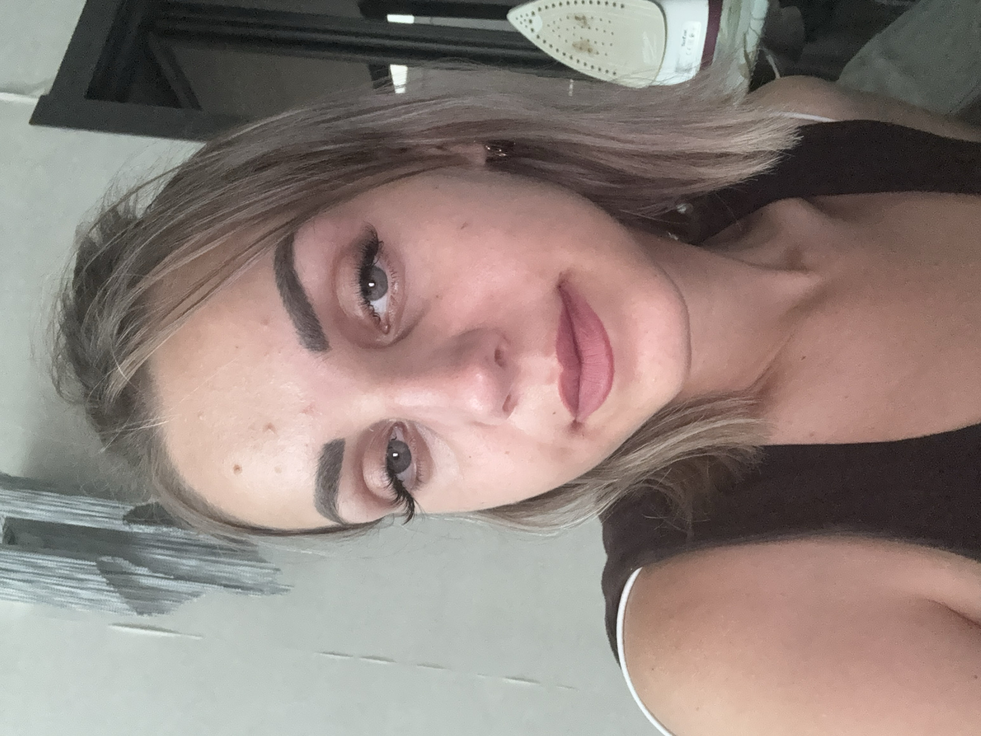 A young woman with blonde hair and makeup poses for a selfie in what appears to be a home setting. She is wearing a dark sleeveless top with a white trim. Behind her, a portion of a wall with peeling wallpaper is visible, and to the right, an ironing board with an iron is partially in frame, suggesting the location might be a room with household appliances. The background also includes a dark doorway. The woman's expression is neutral. The scene does not offer any specific indicators that would suggest a location in Yuzhno-Sakhalinsk, Russia.