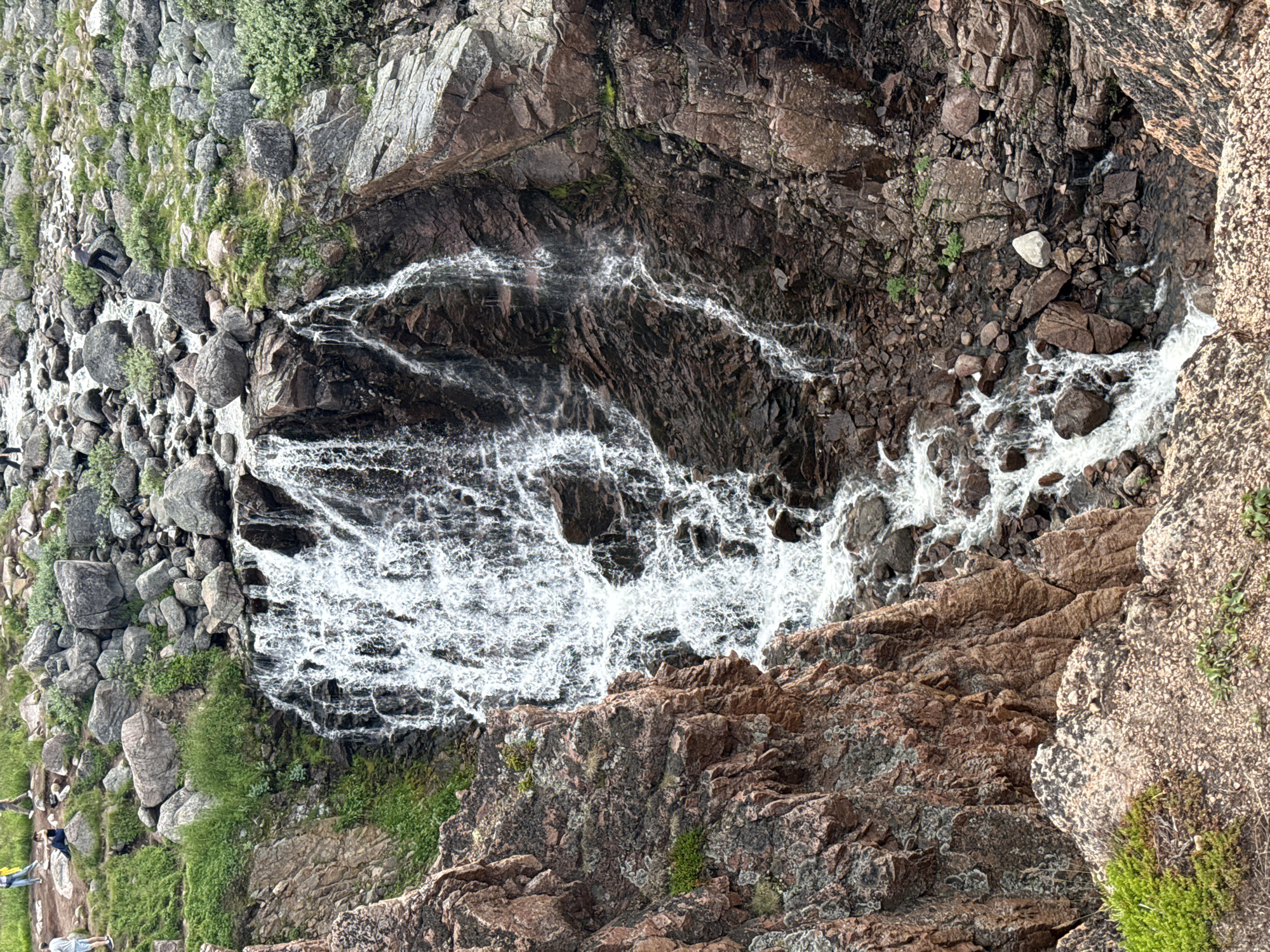 The image depicts a cascading waterfall flowing over a rugged, rocky cliff face in Teriberka, Russia. The waterfall is the central focus, with white water plunging down a steep, dark-colored rock formation. The surrounding environment is characterized by scattered boulders, patches of green vegetation, and more rocky outcrops. In the upper portion of the image, on a flatter, grassy area above the waterfall, several people are visible. One person in a yellow jacket is standing, while others are seated or walking. A person in a grey shirt is also seen navigating the rocks to the right of the upper waterfall. The overall scene conveys a natural, outdoor setting with signs of human presence.