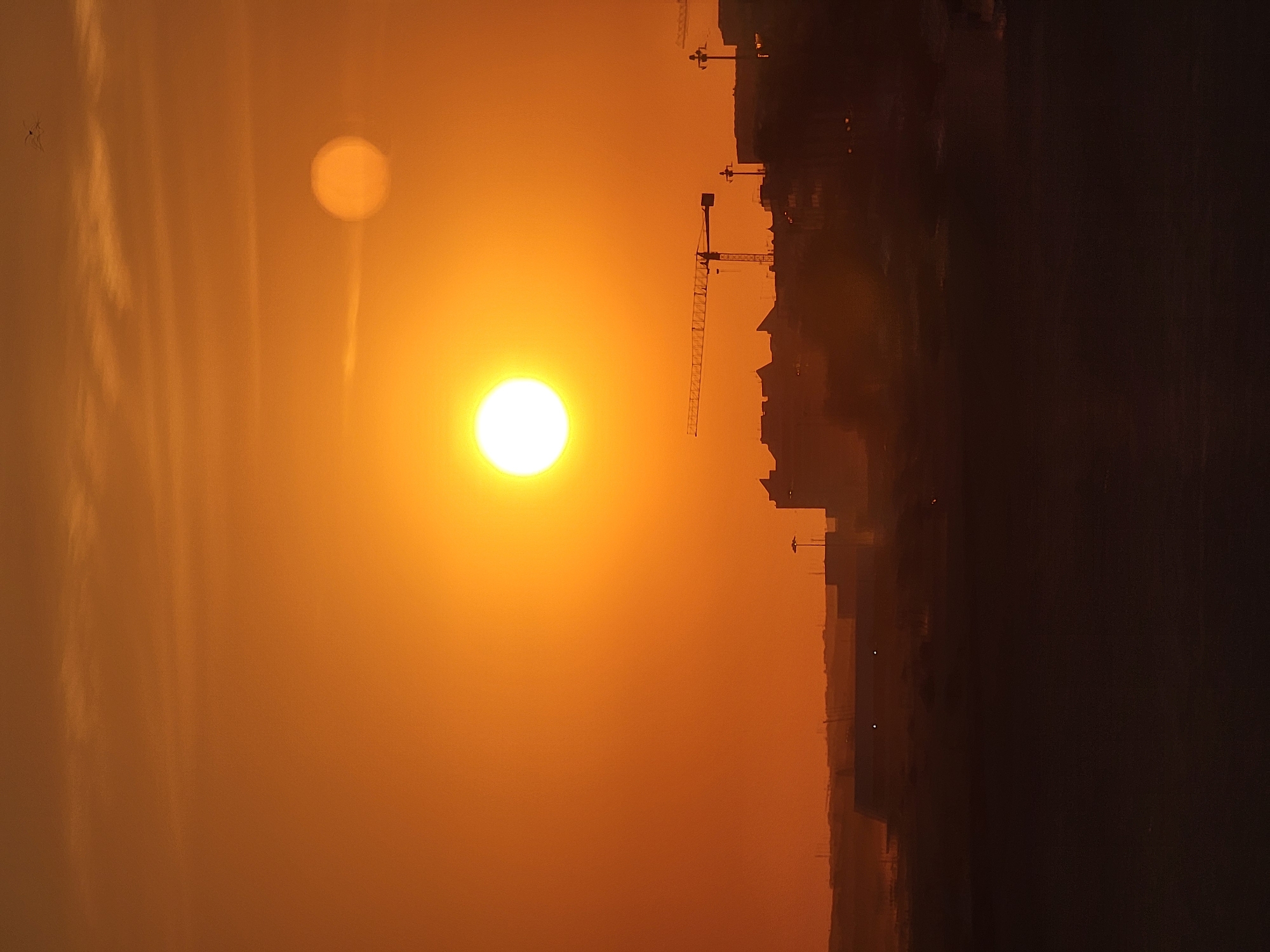 The image depicts a sunset over the city of Quarteira, Portugal. Two prominent suns are visible in the orange sky, with the larger sun centered and a smaller, fainter sun to its right. Silhouetted against the vibrant sky are several buildings and structures, including at least two construction cranes. The foreground is a dark, indistinct mass, likely representing land or water. The scene conveys a serene and atmospheric end to the day.