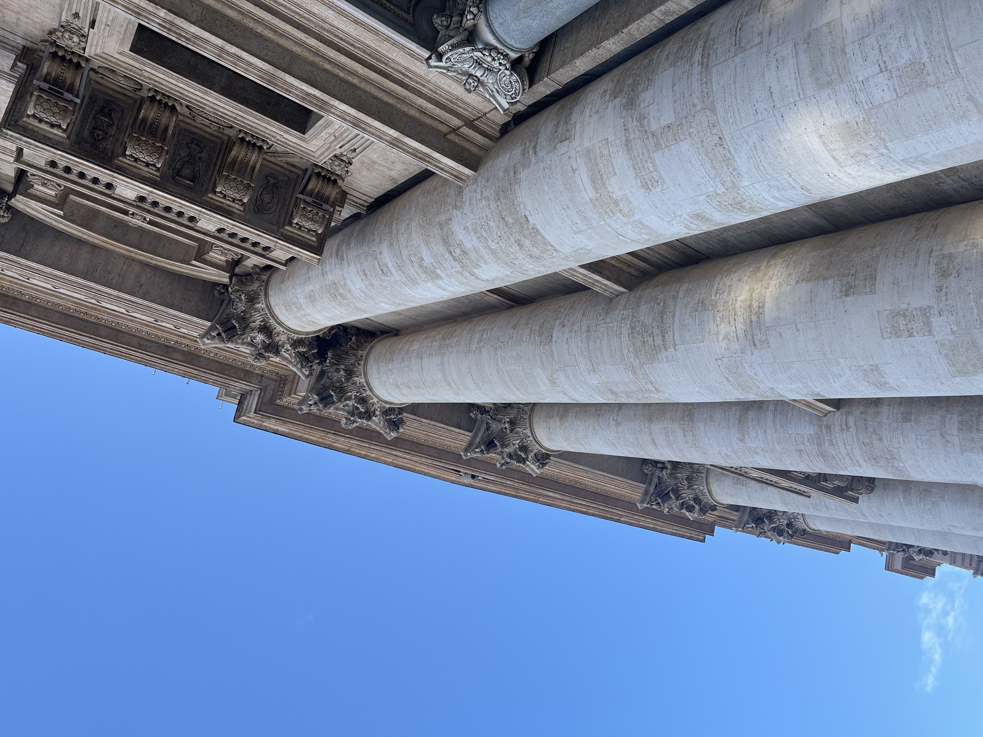 The image displays a low-angle perspective of the monumental stone facade of St. Peter's Basilica in Città del Vaticano, Italy, seen against a clear, vibrant blue sky. Several colossal columns, constructed from light-colored travertine with a visible textured surface, dominate the frame, ascending from the bottom edge towards the top. These columns feature ornate Corinthian or Composite capitals with acanthus leaves and volutes. Above the columns, a grand entablature is visible, comprising multiple layers of highly decorative architectural elements, including intricate friezes, cornices, dentils, and sculpted panels depicting motifs such as urns and foliage. To the far right, partially obscured, a single darker-colored column with a distinctly carved capital featuring putti or angelic figures and scrollwork is visible, set slightly back from the foreground columns. Sunlight illuminates the building, creating subtle shadows that enhance the three-dimensional quality of the architectural details. No people or specific actions are depicted within the frame.