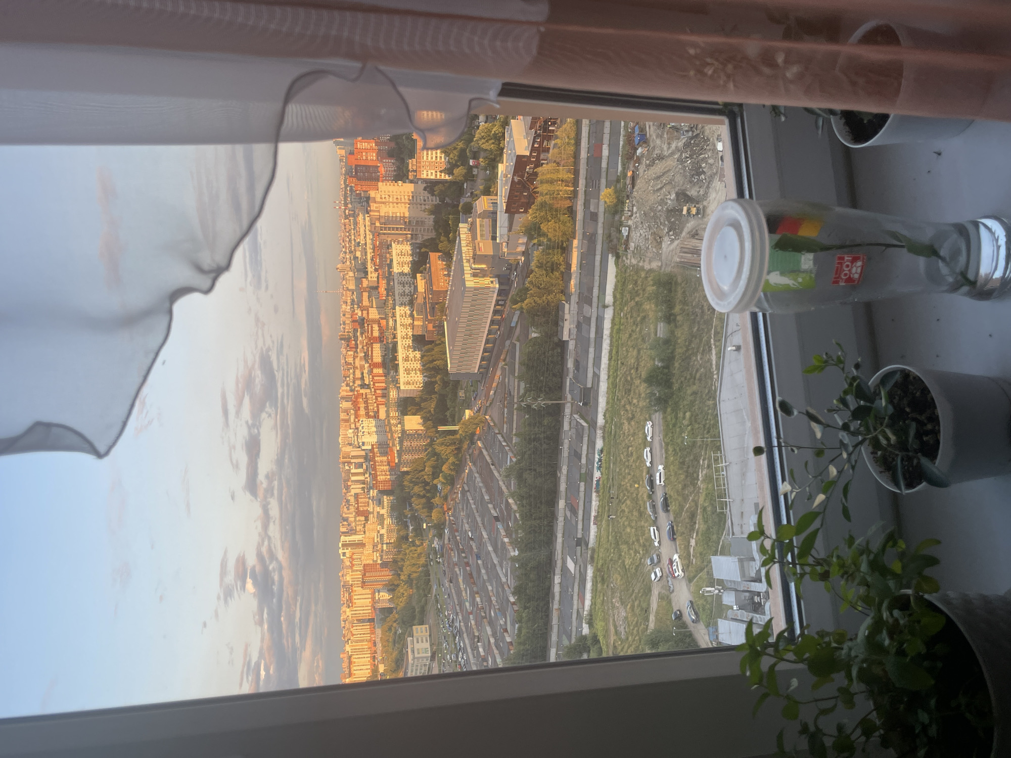 The image, taken from a high-rise window, shows a sprawling city skyline at sunset. The sky is a gradient of soft blues and pinks, with scattered clouds. In the foreground, on the windowsill, are three potted plants, one of which is a small green leafy plant in a white pot. Next to the plants is a clear glass vase with a white lid, containing a single green stem and a label with a red and yellow symbol.
The mid-ground reveals rows of garages or storage units, a parking lot filled with cars, and a modern building with a silver facade. Beyond this, the city extends into a dense urban landscape of apartment buildings, many of which are bathed in the warm glow of the setting sun. Further into the distance, a tall, slender tower is visible on the horizon. The overall environment appears to be a residential area within a city, with a mix of old and new architecture. A construction site with a pile of rubble is visible in the lower right quadrant of the image.
