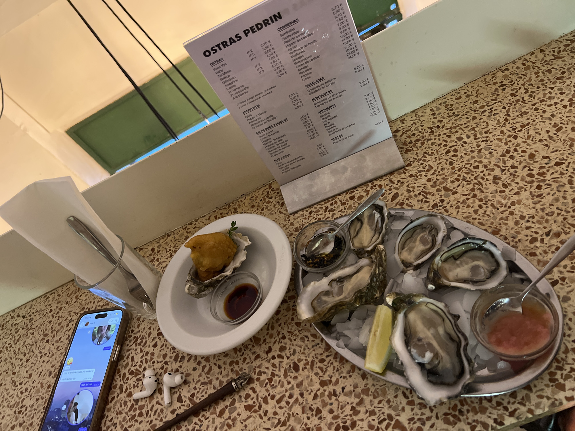 Here is a forensic description of the image:
**Location:** València, Spain, presumably at a restaurant named "Ostras Pedrin" based on the menu.
**Scene:** A table setting featuring an assortment of oysters, sauces, and other items.
**Objects:**
* An iPhone, displaying a chat interface on its screen.
* A pair of wireless earbuds.
* A pen.
* A menu for "Ostras Pedrin," listing various seafood dishes and prices.
* A glass containing napkins and silverware.
* A white dish containing two oysters, one with a fried topping. A small dish with soy sauce is also on this plate.
* A metal tray holding several fresh oysters on ice.
* Two small bowls with dipping sauces.
* A lemon wedge.
* A spoon is on each oyster platter.
* Terrazzo table.
**Food:** The focus of the image is on seafood. There are oysters, both fresh and in a prepared style (likely tempura or fried). The accompanying sauces appear to be various condiments for the oysters.
**Environment:** The table is set against a backdrop of what appears to be the interior of a restaurant. The tabletop has a terrazzo pattern. The area is well-lit.
**Summary:** The image depicts a restaurant setting where a person is about to enjoy a meal of oysters and possibly other seafood dishes. The presence of a phone, earbuds, and pen suggests the person may be engaged in communication or work-related activities while dining.