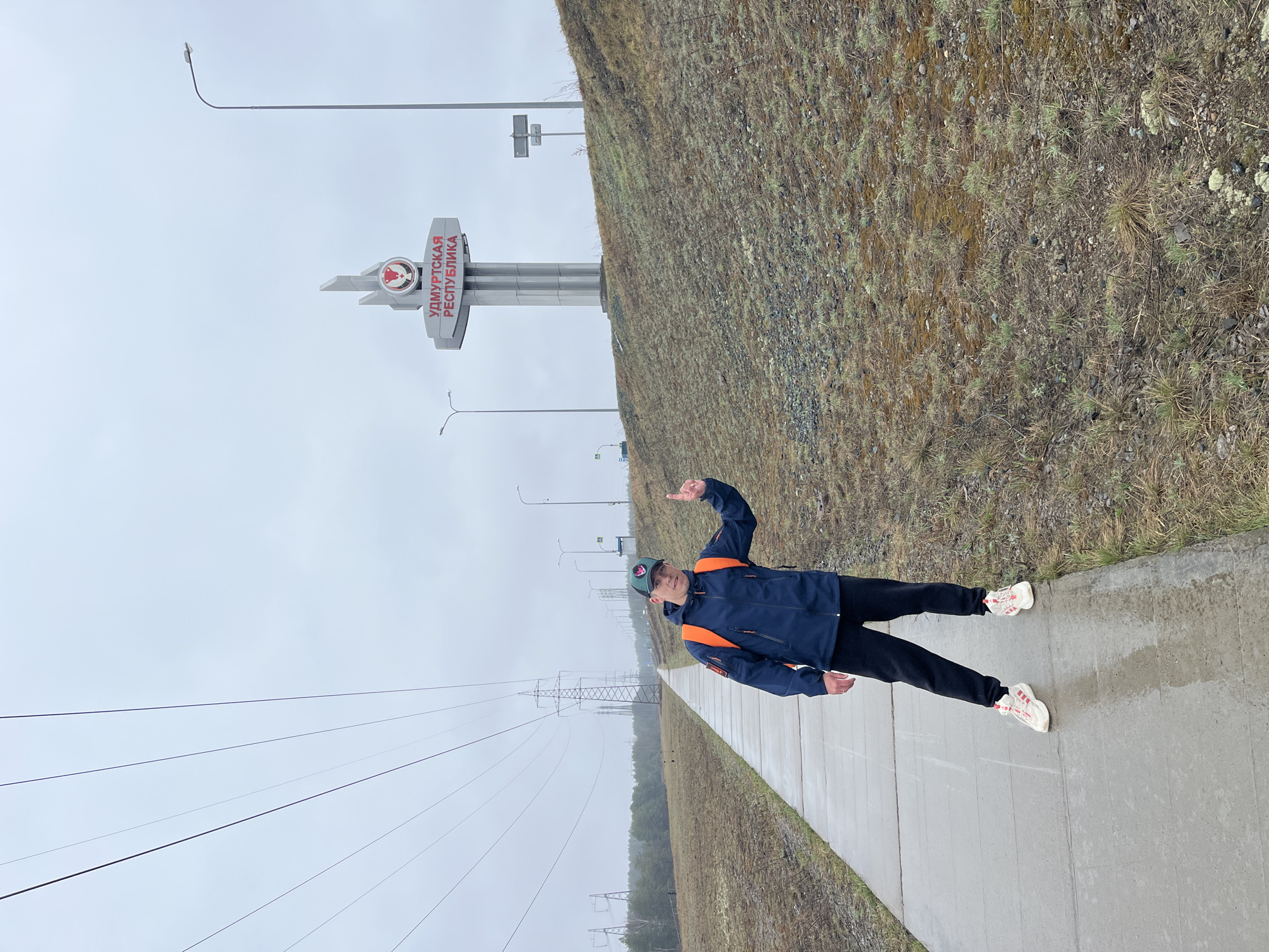 A full-body shot captures a man posing on a wet concrete path beside a roadside monument under an overcast sky. He is dressed in a dark blue jacket with bright orange accents, black pants, and light-colored sneakers with red details, topped with a baseball cap. He faces the camera, pointing upwards with his right index finger and giving a thumbs-up with his left hand, indicating a positive and perhaps celebratory mood.
Behind the man is a prominent grey monument featuring a red emblem, possibly an elk or moose head, above the Cyrillic text "УДМУРТСКАЯ РЕСПУБЛИКА" (Udmurt Republic). This sign likely marks an administrative border crossing into the Udmurt Republic, consistent with the provided location context of Chaikovsky, Russia, which is geographically close to this region.
To the man's right, a gently sloping hill covered in sparse, dry grass and some small rocks rises. To his left, multiple power lines stretch across the frame, supported by distant power pylons. Streetlights are also visible along the path extending into the background. The ground, including the concrete path, appears damp, suggesting recent rain or high humidity. The sky is uniformly grey and overcast, contributing to a cool, subdued atmosphere.