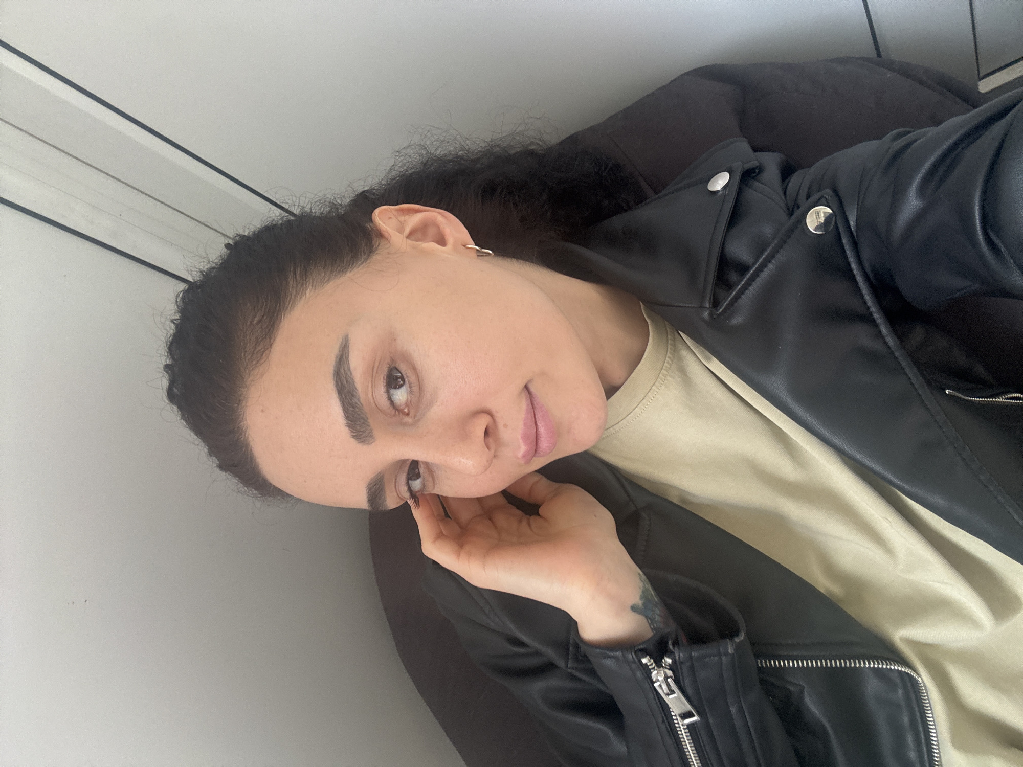 A woman with dark curly hair is posing for a selfie. She is wearing a tan-colored t-shirt and a black leather jacket. Her left hand is resting on her cheek. The background consists of a white wall with black and gray accents. There is no information available to determine the specific location within Tbilisi, Georgia.