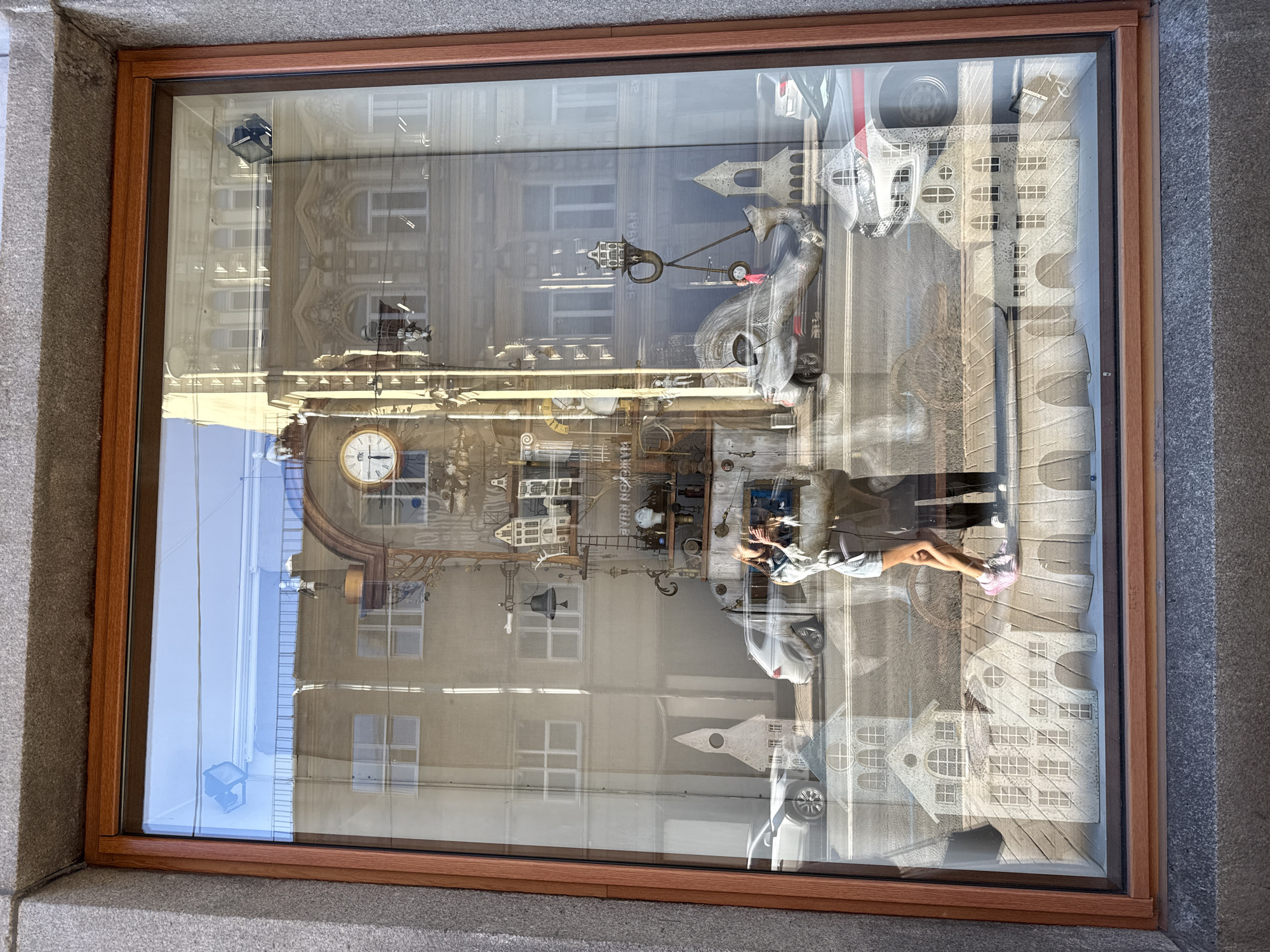 This image shows a street-level window display, likely in Saint Petersburg, Russia. The display is a diorama featuring miniature buildings and structures, possibly representing a street scene. The glass reflects the sky and buildings opposite the display, creating a complex layered image.
The diorama itself includes several elements. In the upper left, a clock face is visible, suggesting a town square or historic district. Various mechanical and industrial-looking objects, along with miniature architectural details, are arranged throughout the display. These include what appear to be cranes, gears, and scaffolding, integrated with small building facades.
A prominent feature is a mannequin or doll lying down, wearing a light-colored shirt and pants, with pink sneakers. The figure is positioned on a surface that resembles a wooden deck or walkway within the diorama. The reflection of the building opposite the window is strong, showing its architectural details, windows, and balconies. The lighting suggests it is daytime, possibly late morning or early afternoon, with bright sunlight casting distinct shadows. The sky appears clear, indicating fair weather.
There is no visible text in the image. The overall impression is a whimsical, perhaps steampunk-inspired, miniature cityscape integrated with the reflections of the real world outside the window.