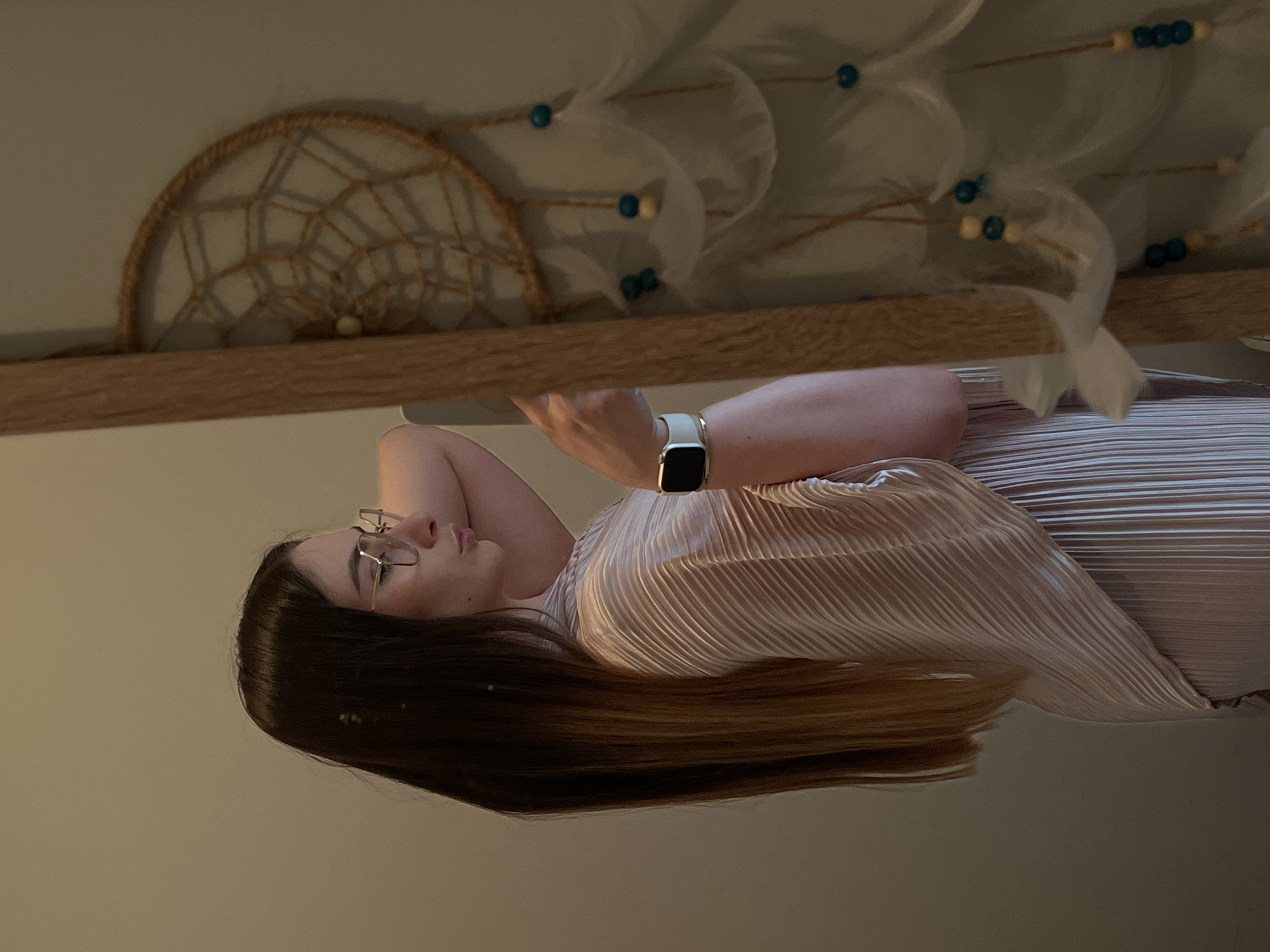 A woman is lying on her side, her head resting on her arm. She is wearing glasses and a smart watch. Her long hair is mostly dark brown, transitioning to a lighter shade at the ends. She is wearing a light pink pleated top. Behind her is a wooden shelf with a dream catcher and other decorative items with beads and white feathers. The background is a light beige color. The location is Unknown, Unknown.