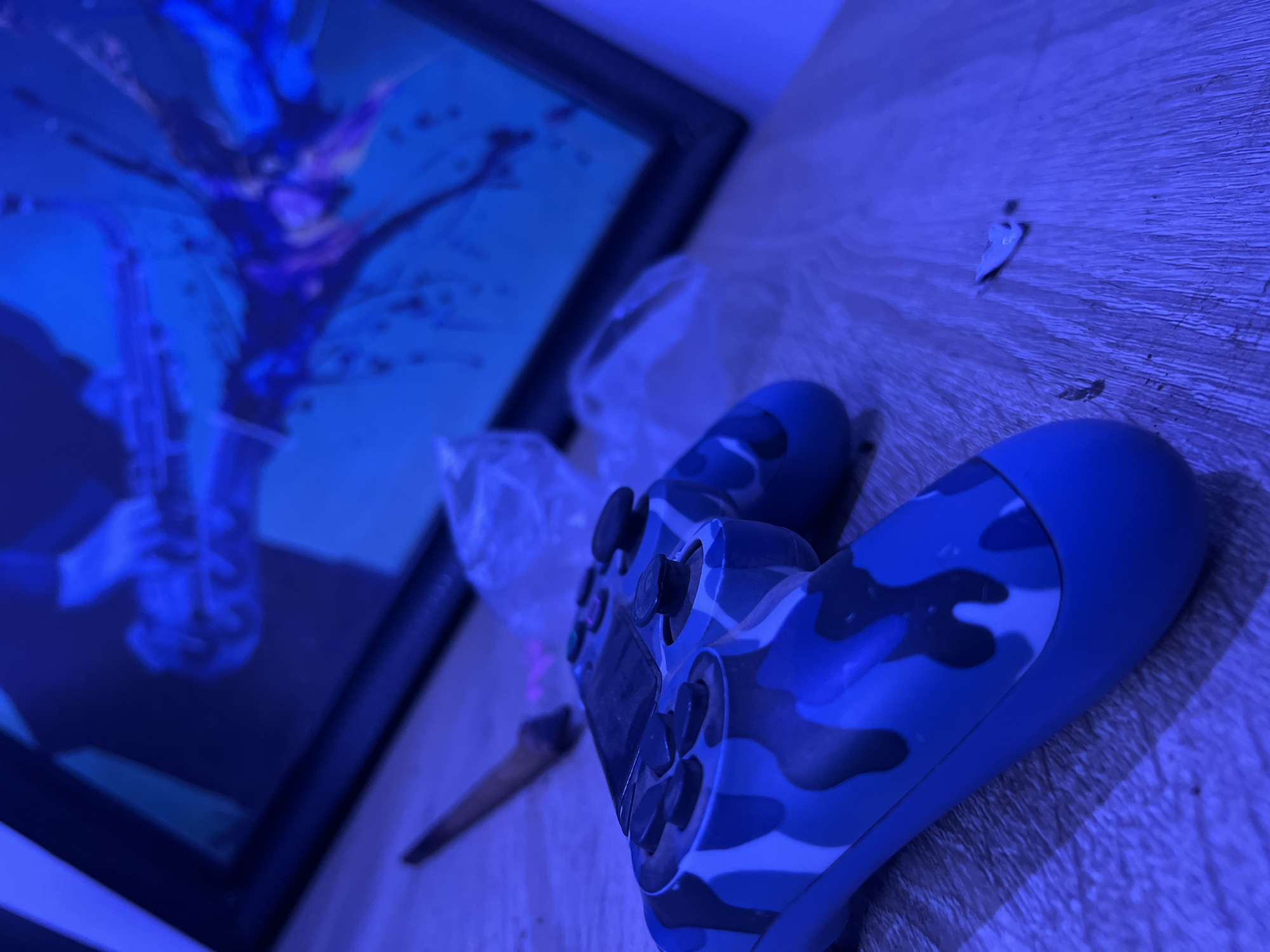 A blue camo PlayStation 4 controller rests on a wood-grain surface, partially obscured by translucent plastic bags and a dark, thin object resembling a rolled cigarette. A framed artwork depicting a saxophone player in blue hues is visible in the background. A small piece of paper or debris lies on the wood-grain surface near the controller. The scene is bathed in blue light. No people are visible. The specific location in Abuja, Nigeria cannot be determined from the provided image.