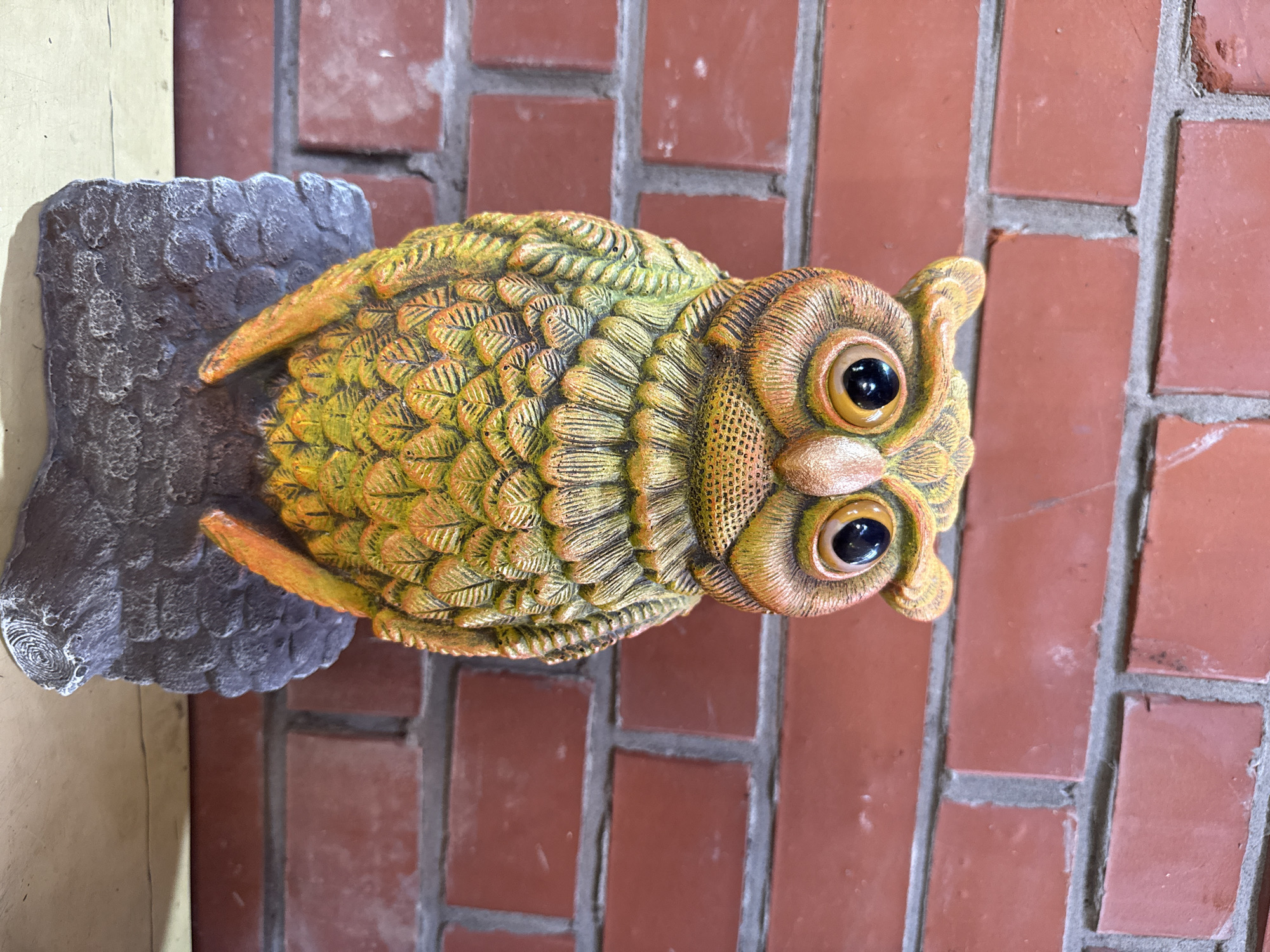 A close-up, eye-level shot shows a decorative ceramic owl figurine positioned in front of a red brick wall. The owl is depicted with its head tilted slightly to its left, displaying large, dark, reflective eyes and an orange-brown face with textured details resembling feathers. Its body is primarily a gradient of green and yellow, with intricate feather patterns painted onto its surface. The owl is perched on a dark gray, rocky base with a textured, rough finish. The brick wall behind the owl is composed of rectangular red bricks with visible gray mortar lines. The lighting appears to be ambient, casting subtle shadows. The scene is static and devoid of human presence or activity.