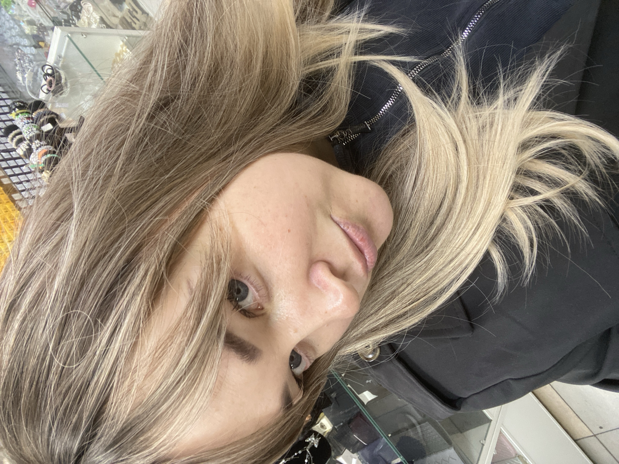 A close-up portrait, oriented at a tilt, features a young woman with long, flowing hair that transitions from a light brown base to blonde highlights. Her head is angled, with her hair draped around her face and shoulders, resting against a black jacket or top with a visible silver zipper.
The woman's right eye, part of her nose, and her lips are visible, with her gaze directed slightly to the upper left of the frame (our right). Her skin appears clear, showing a few natural freckles on her nose and cheek. Her lips have a subtle pink hue.
In the blurred background, several glass display cases filled with various jewelry and accessories are visible. These include rings, earrings, and other shiny, decorative items, some appearing to be silver-toned, like a bow-shaped pendant. A grid-like pattern on a surface, possibly a display stand, can also be seen. The setting suggests an indoor commercial space, likely a jewelry or accessories shop.
The woman appears to be posing for a photograph or selfie. No specific time of day or weather can be determined from the image, nor is any readable text visible.