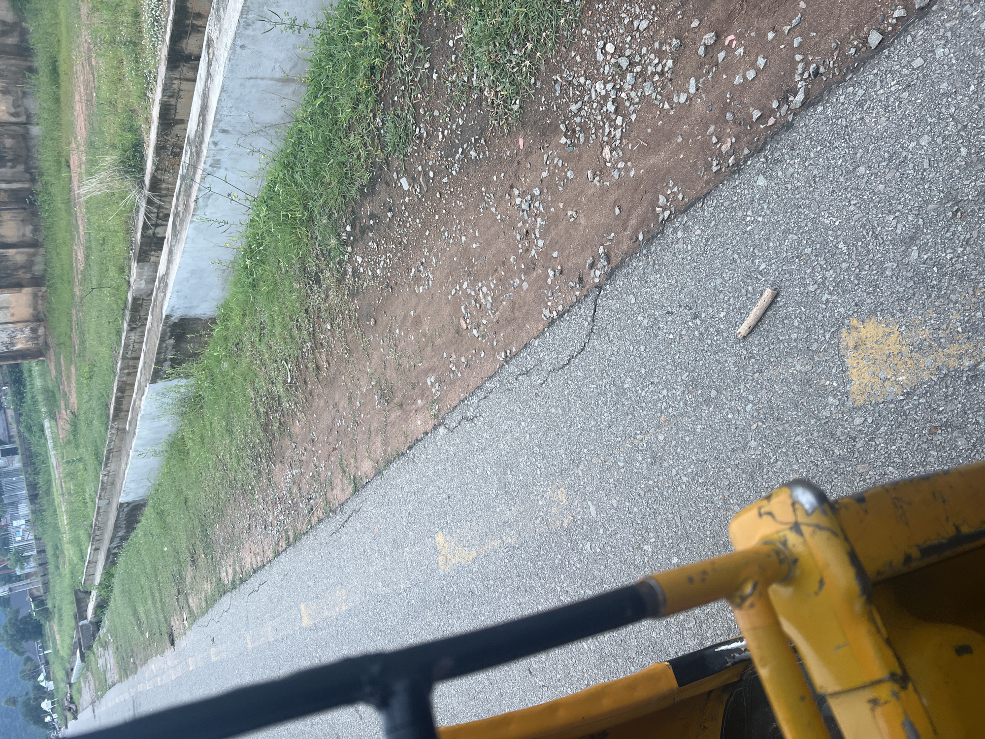 The image depicts an outdoor scene, likely taken from a vehicle, given the perspective and the presence of yellow and black railings in the foreground. The setting appears to be a roadside adjacent to a grassy embankment and a concrete structure, possibly a drainage channel or a retaining wall. The ground surface is a mix of asphalt with yellow markings and a dirt path scattered with small stones and pebbles. Sparse green grass grows along the edge of the embankment.
In the distance, on the left side of the frame, there are glimpses of buildings, suggesting an urban or semi-urban environment. The sky is not visible, but the lighting suggests it is daytime, possibly overcast or early morning/late afternoon. There are no people visible in the image.
A single, thin twig lies on the asphalt surface, a small detail in the overall scene. The image is oriented vertically, which may be due to the camera's orientation or the nature of the photograph. The overall impression is one of a functional, somewhat utilitarian environment, perhaps a construction site or an industrial area given the presence of heavy machinery implied by the yellow railings. The location context provided, Kona Garu, Nigeria, places the scene in a specific geographical region.