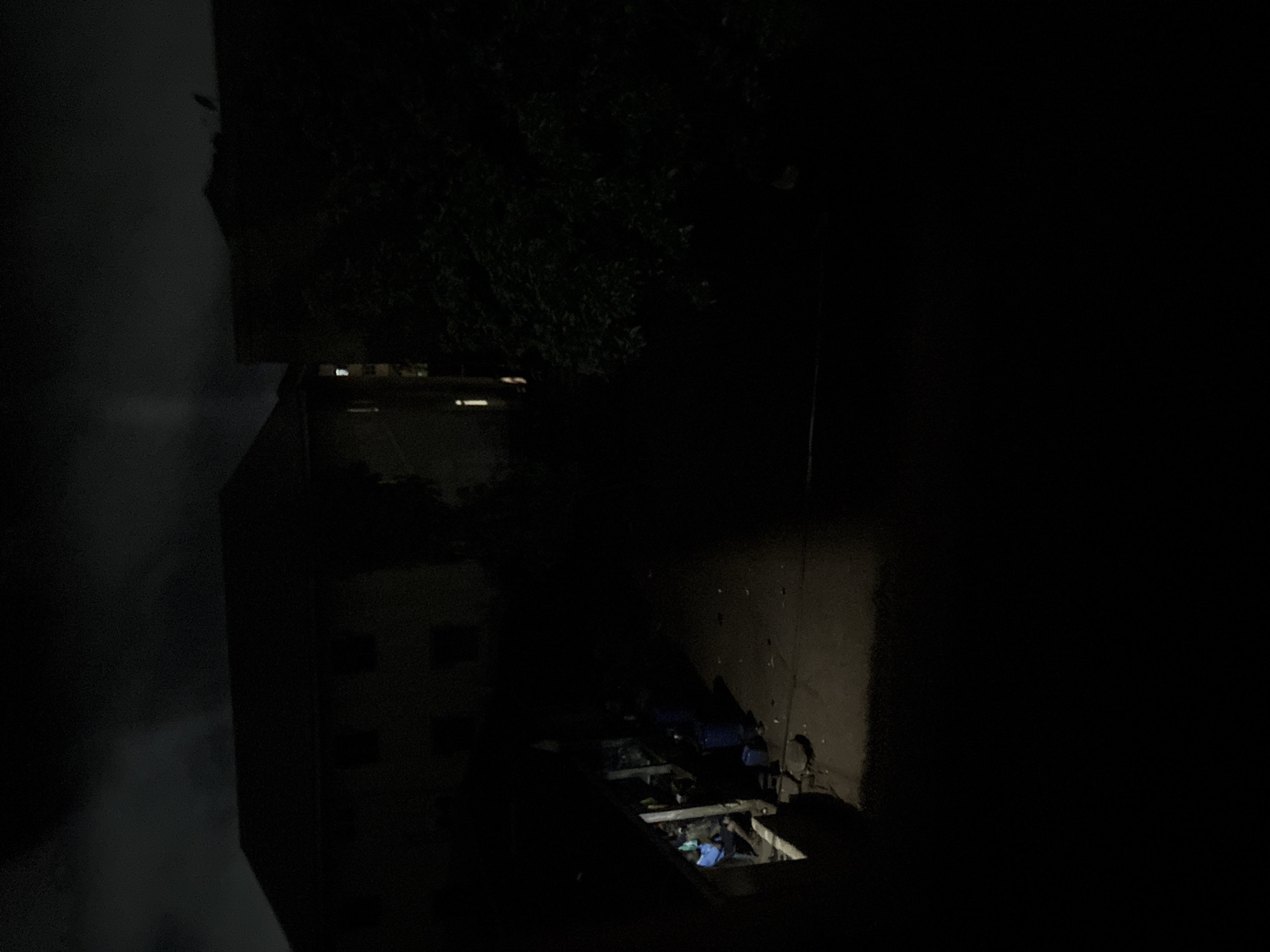 The image depicts a dimly lit outdoor scene at night, possibly in a residential area of Onitsha, Nigeria. The foreground is largely obscured by darkness. In the mid-ground, a building with a dark roof and visible windows is present. One window on the left side of this building is illuminated from within, suggesting occupancy. To the left of this building, there is an open-air structure, possibly a shelter or workshop, where two individuals are visible. One person, wearing a blue garment, appears to be interacting with materials or objects on a surface, while another person is situated nearby. Two blue plastic containers are also present in this area. A dark, dense tree or shrubbery is located to the right of the building. The ground surface appears to be paved or dirt. The sky above is dark with some faint cloud formations. The overall impression is a snapshot of nighttime activity in an urban or semi-urban environment.