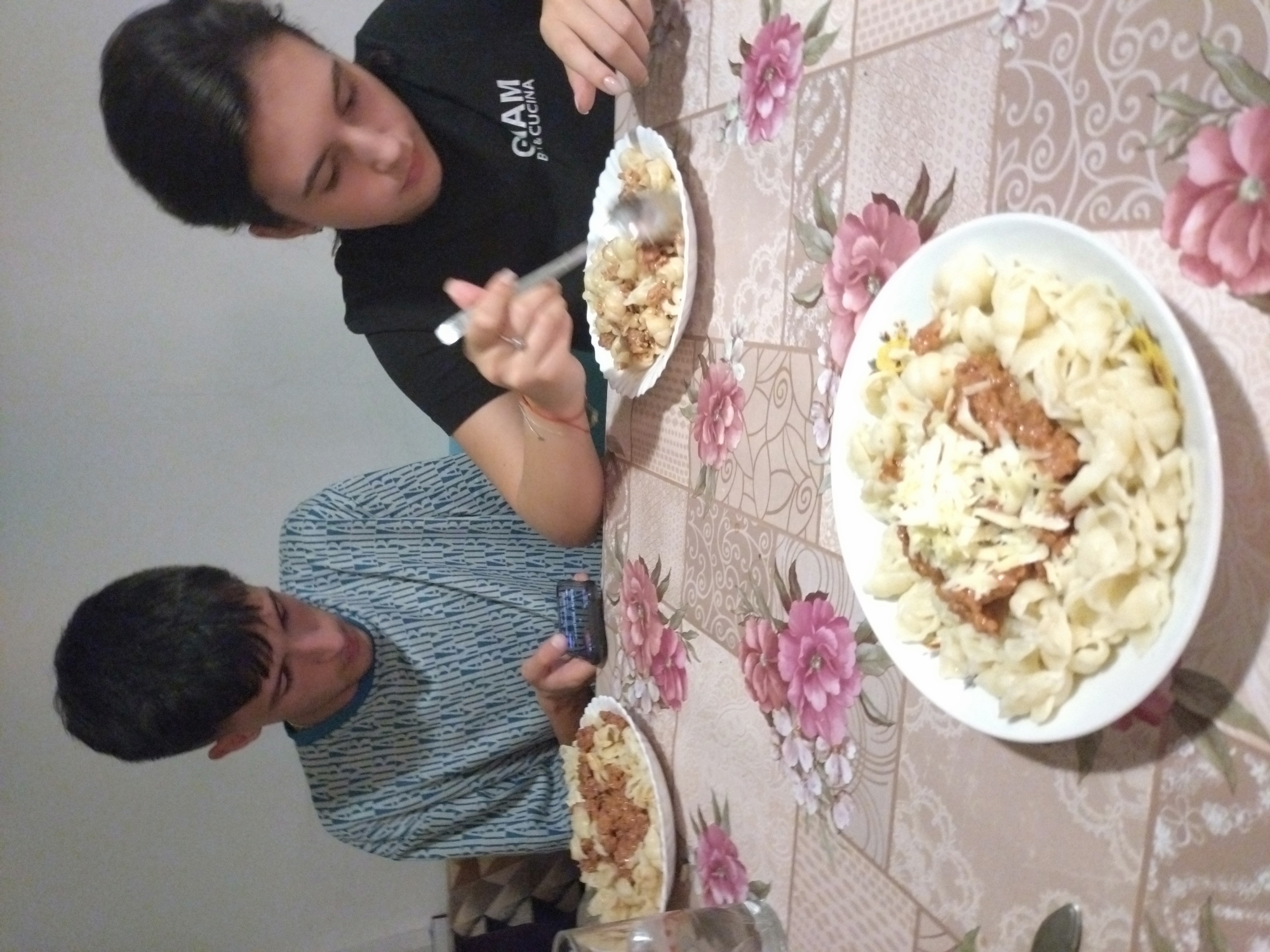 Two individuals are seated at a table covered with a floral patterned tablecloth. The person on the left is holding a smartphone and has a plate of pasta with meat sauce in front of them. The person on the right is holding a fork and eating from a similar plate. A larger bowl of pasta with what appears to be meat sauce and grated cheese sits on the table as well. The scene is indoors. There is no definitive evidence within the image to confirm that the location is Stara Zagora, Bulgaria.