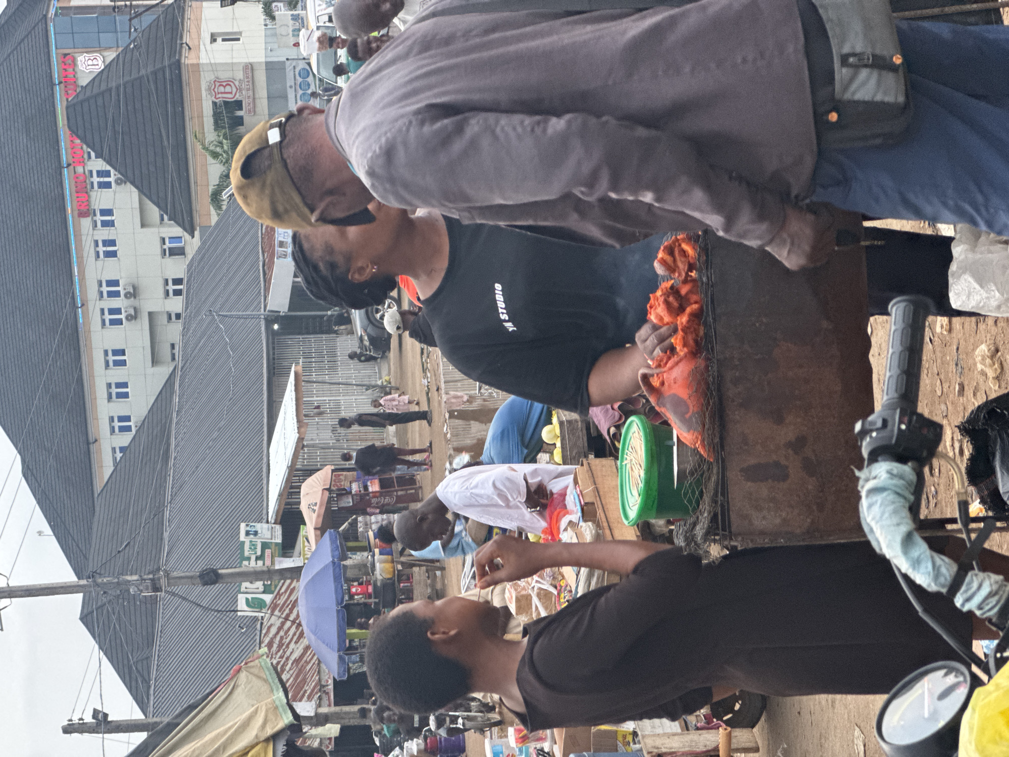 The image captures a street scene in Obinze, Nigeria, likely a market area, under an overcast sky. In the foreground, a woman in a dark dress stands facing a grill with pieces of cooked chicken. To her right, a man in a grey jacket and blue pants is talking to a woman wearing a black t-shirt with "YN STUDIO" printed on it. The grill, a rudimentary metal structure, holds several pieces of what appear to be marinated and grilled chicken. Behind the main subjects, stalls and structures indicative of a market are visible, including a blue umbrella, various goods on display, and other individuals. In the background, modern buildings with glass windows and prominent signage, one reading "BRUNO HOTELS & SUITES," suggest a mix of commerce and accommodation. A motorcycle is partially visible in the lower left corner. The ground appears to be a dirt or gravel surface.