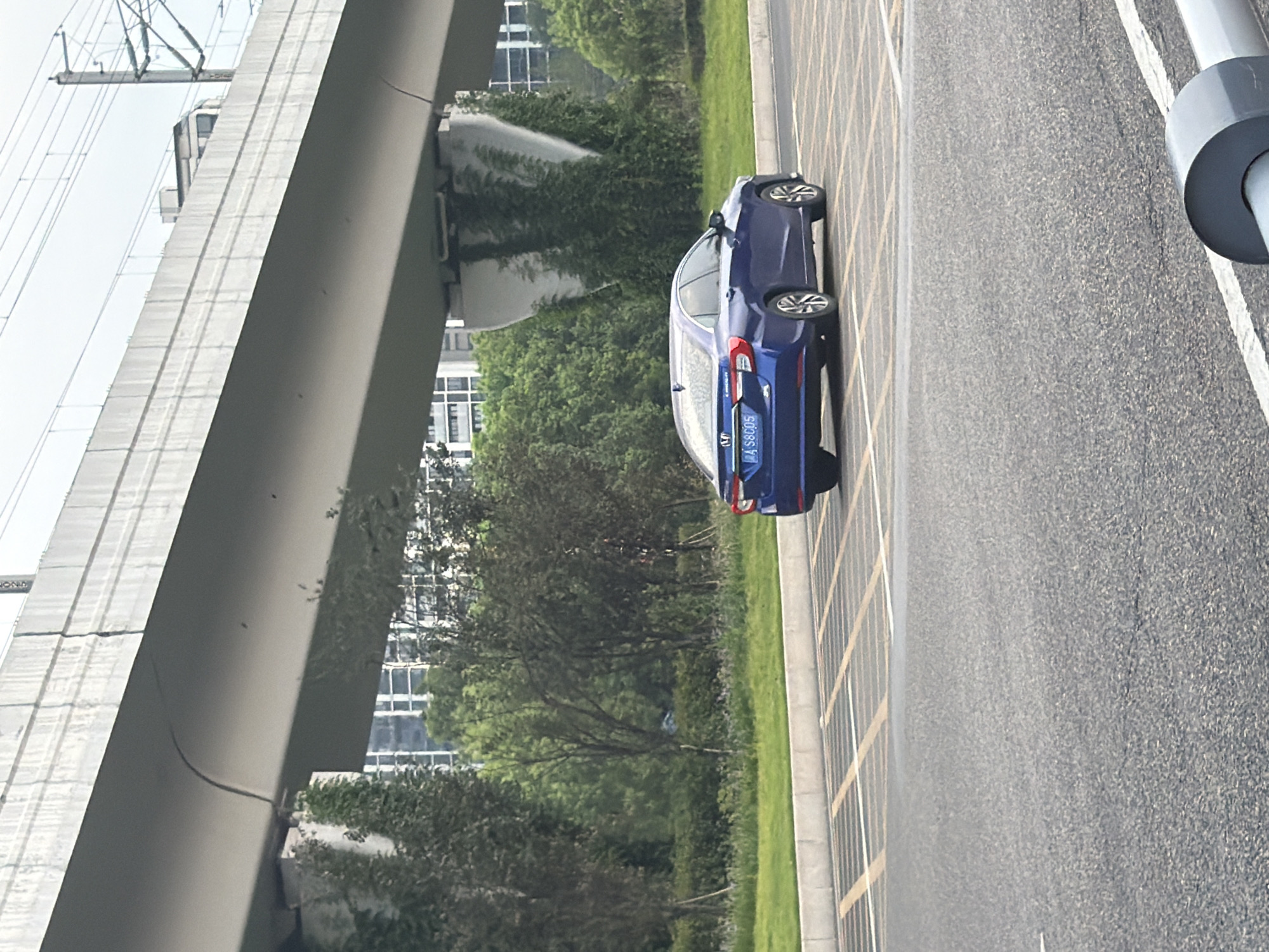 A blue sedan, license plate "A S8C05", is parked on an asphalt surface marked with yellow and white lines, possibly a parking lot. The car is positioned at an angle, facing towards the right of the frame, with its rear visible. Behind the car, a patch of green grass meets a line of trees and further back, several tall buildings rise against a cloudy sky. An elevated structure, likely a bridge or overpass, dominates the upper portion of the image, supported by concrete pillars overgrown with vines. The scene appears to be in an urban or suburban area in Jiangjin, China, with clear evidence of infrastructure like the elevated roadway and modern buildings.