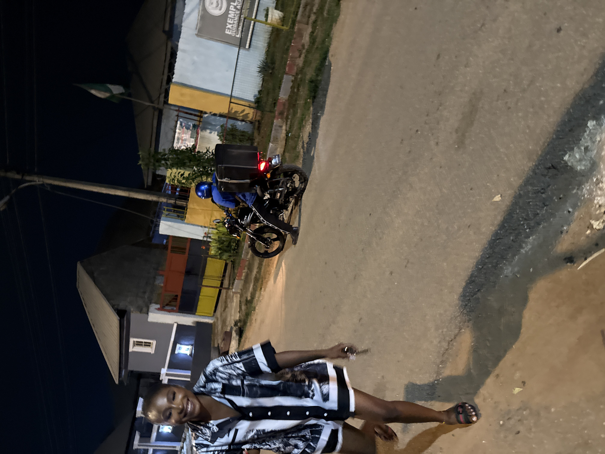 In an urban street scene at night in Abuja, Nigeria, a young woman with short hair and a radiant smile is captured in the foreground, walking towards the camera. She is wearing a black and white patterned short-sleeved shirt and shorts. In the background, a person wearing a blue helmet and jacket is riding a motorcycle with a black cargo box attached to the rear. The motorcycle has its taillight illuminated. The environment appears to be a residential or commercial area with buildings visible on either side of the street. One building features a prominent Nigerian flag. The road is paved and appears to be in moderate condition, with some cracks and debris. The lighting suggests it is nighttime, with artificial lights illuminating the scene.