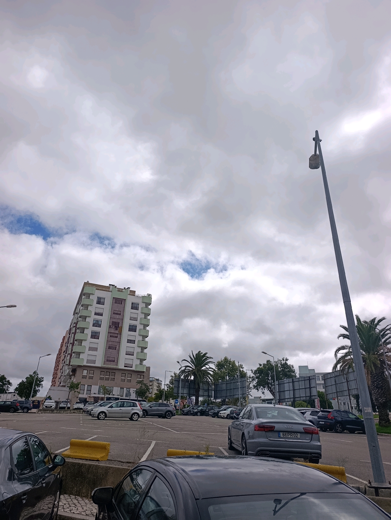 The image captures a daytime scene in a parking lot located in Setúbal, Portugal, near the waterfront or a similar urban area, indicated by the presence of a large apartment building and palm trees. The sky is overcast with scattered white clouds. In the foreground, the rear ends of two dark-colored cars are visible, parked in a lot with yellow concrete barriers. Further into the lot, a silver Audi sedan with the license plate "66 PR02" is parked. Behind it, several other vehicles are visible, including SUVs and sedans. A tall lamppost stands to the right of the frame, and a distinctive, tall apartment building with a light-colored facade and green balconies dominates the mid-ground. Palm trees are scattered throughout the scene, adding to the Mediterranean feel. The overall environment appears to be a typical urban parking area, likely associated with residential or commercial buildings. No people are depicted in the image, and no actions or interactions are taking place.