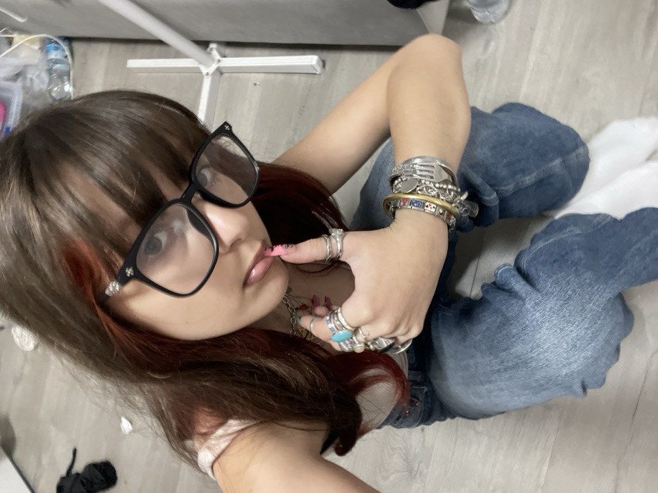 A young woman with long brown hair and bangs wears oversized black glasses. She has a red streak in her hair. She wears several rings on her fingers and numerous bracelets on her wrist. She has on light blue jeans and white socks. She is likely sitting on the floor. Her index finger is touching her lips.