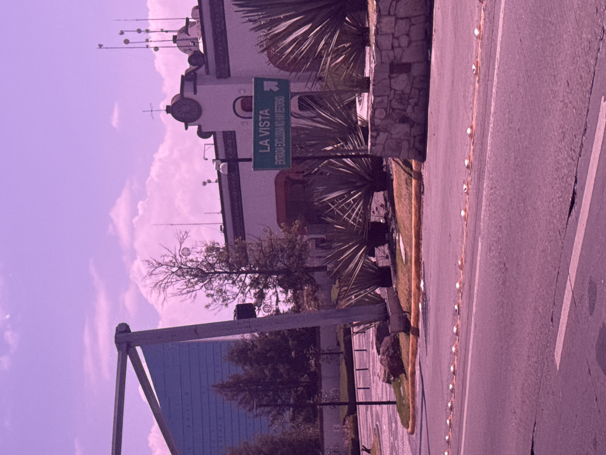 The image depicts a street scene in San Bernardino Tlaxcalancingo, Mexico. A road with reflectors on the edge is visible on the right. A building with a stone facade and multiple antennas on the roof is present. A green sign reading "La Vista" with a right-pointing arrow is mounted near the building. Trees and other vegetation are also visible. A large blue billboard is situated in the lower left corner. The sky is a light purple color.